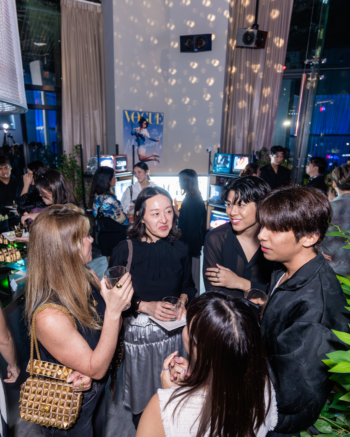 Group of people socializing at a lively party or event with vintage television screens and Vogue magazine poster in the background.