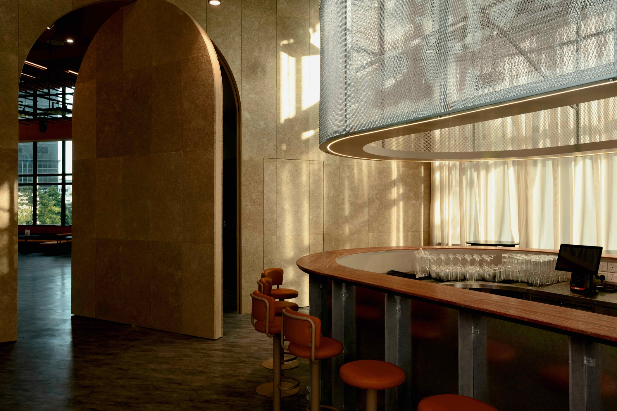 Modern bar with a curved wooden counter, orange bar stools, glassware, and soft lighting, located inside a high-rise building with large windows and beige walls.