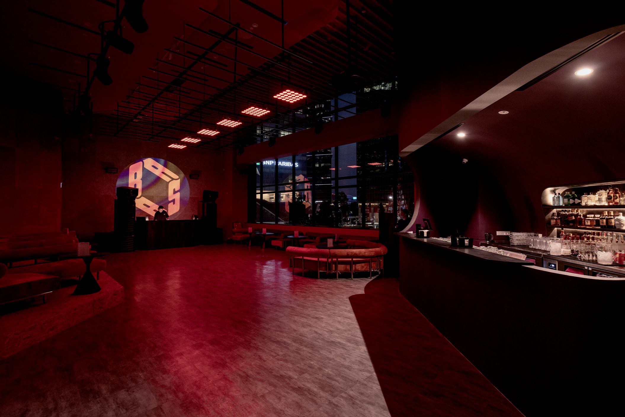 Empty nightclub with red lighting, a DJ booth with a DJ, a bar area, seating along the window, and city lights outside.