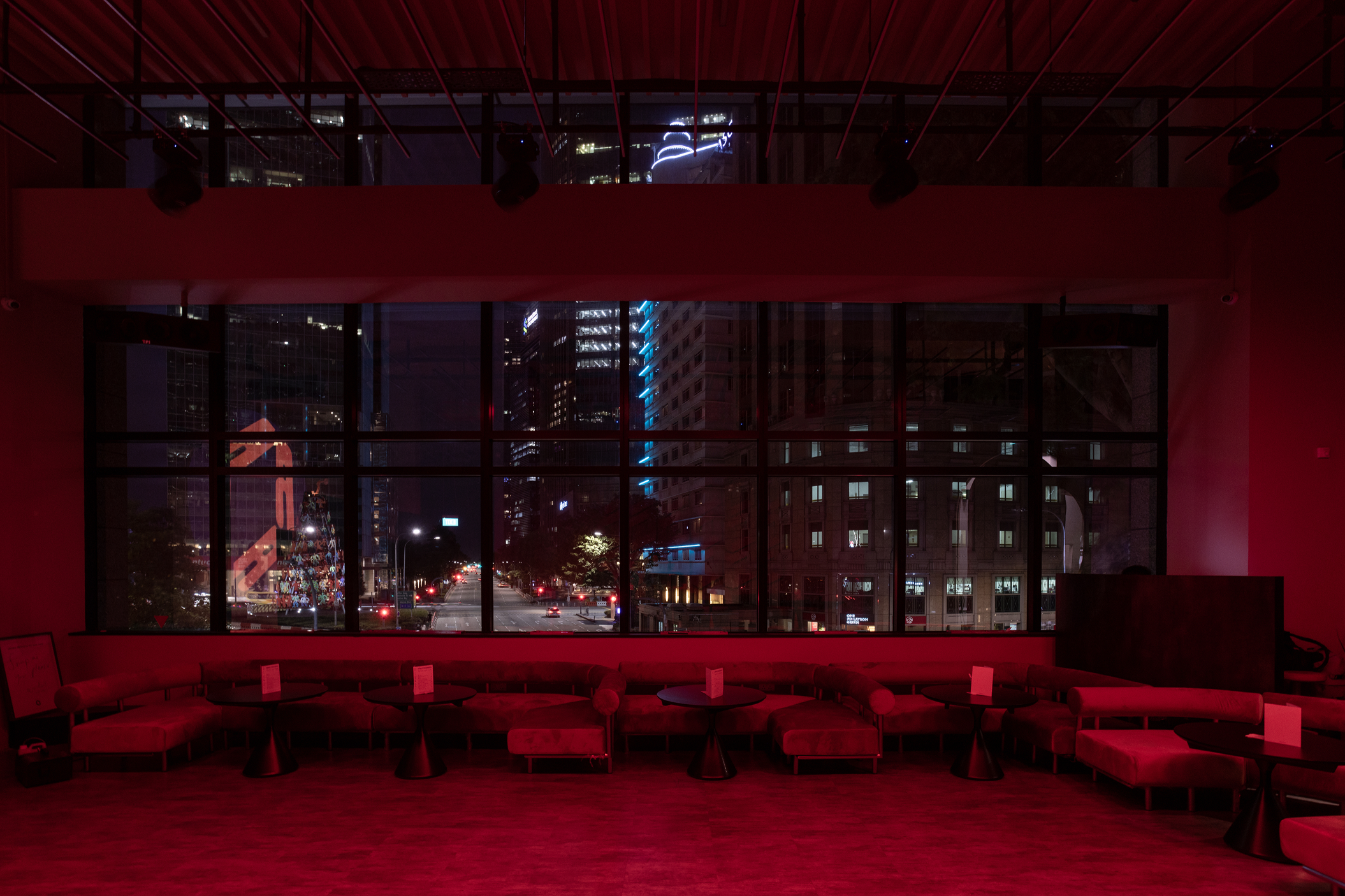 View of a city street at night through large windows, with lit skyscrapers and traffic lights; interior room with red lighting, modern sofas, and small round tables.