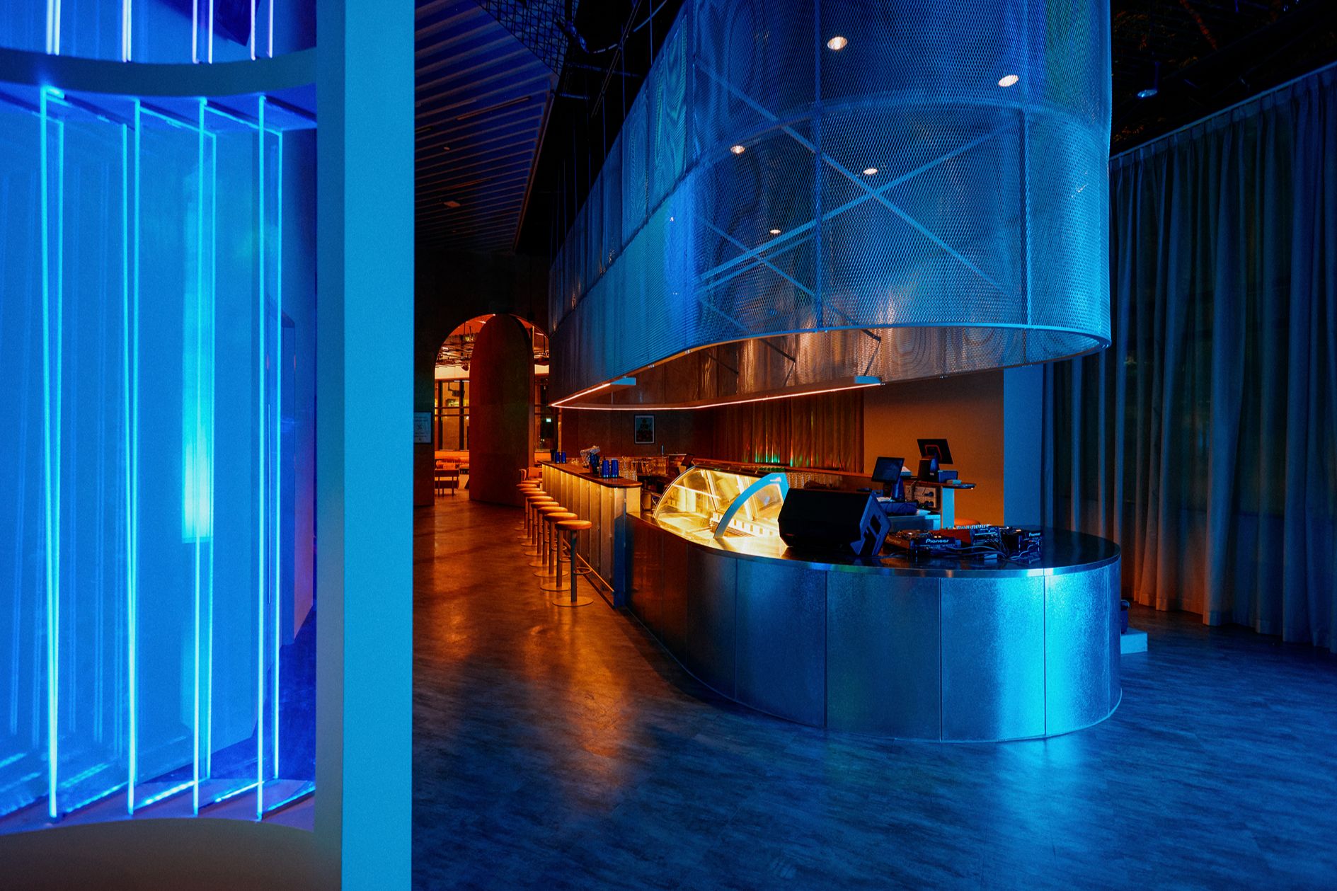Interior of a modern bar with blue and orange lighting, curved metallic counter, and bar stools