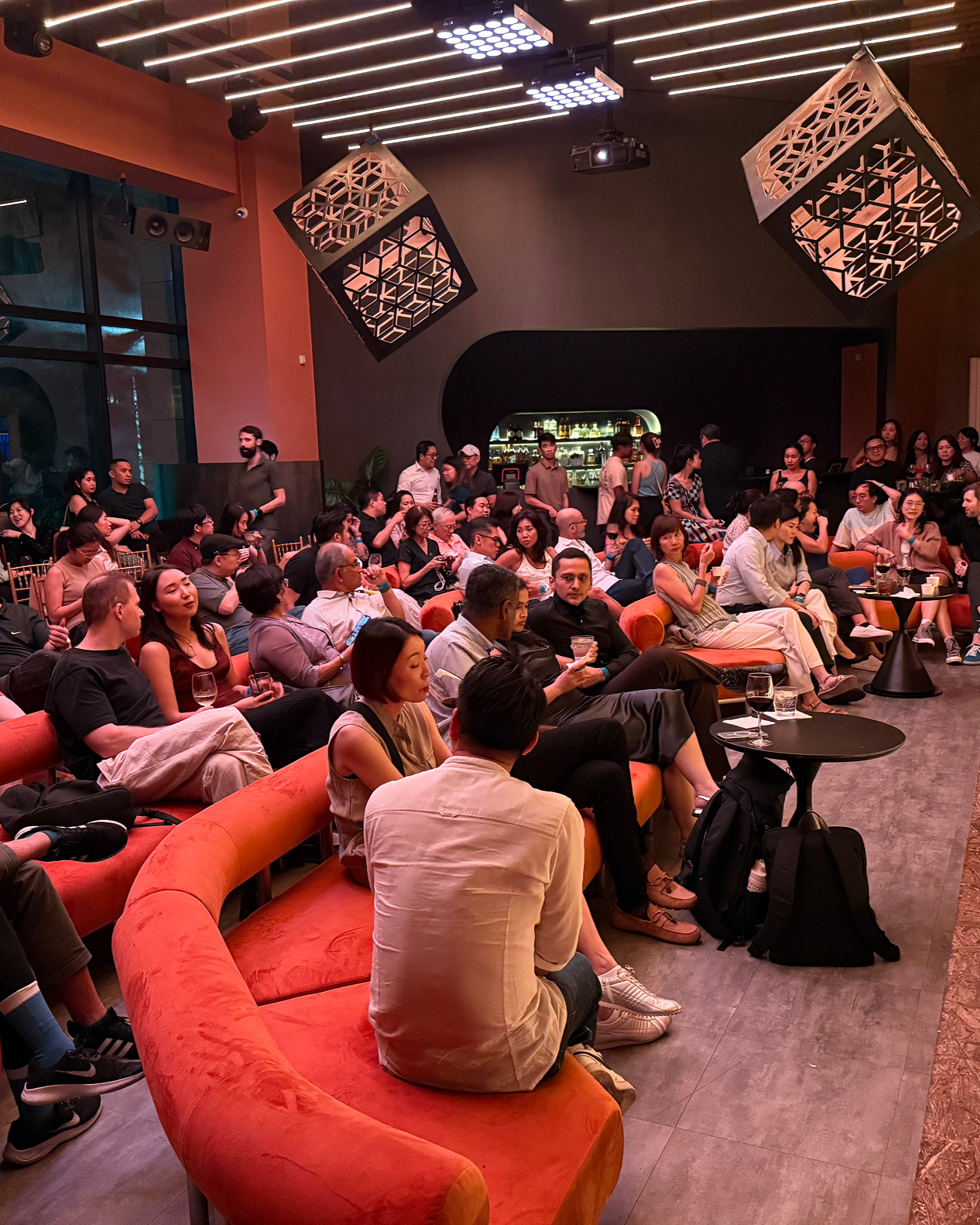 People sitting on orange sofas and chairs watching a performance in a modern, dimly-lit venue with unique hanging light fixtures and a bar in the background.