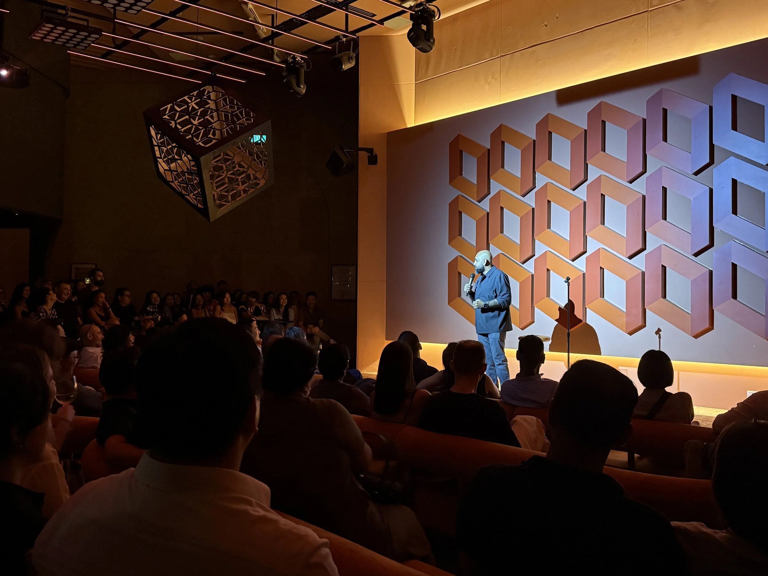 A speaker on stage at a conference or event speaking into a microphone, facing an audience seated in a darkened room with a geometric background design on the wall.