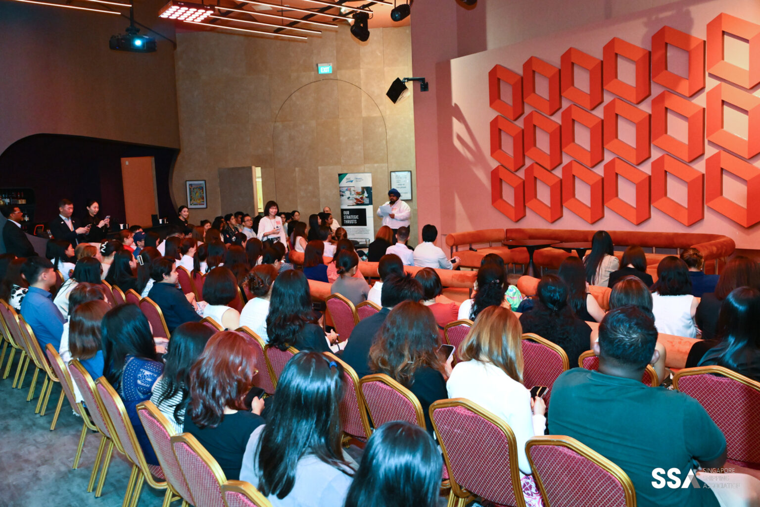 A large audience seated in a conference room, listening to a speaker at the front. The room features a modern decor with a geometric wall art installation, a stage or presentation area, and people taking photos or notes.