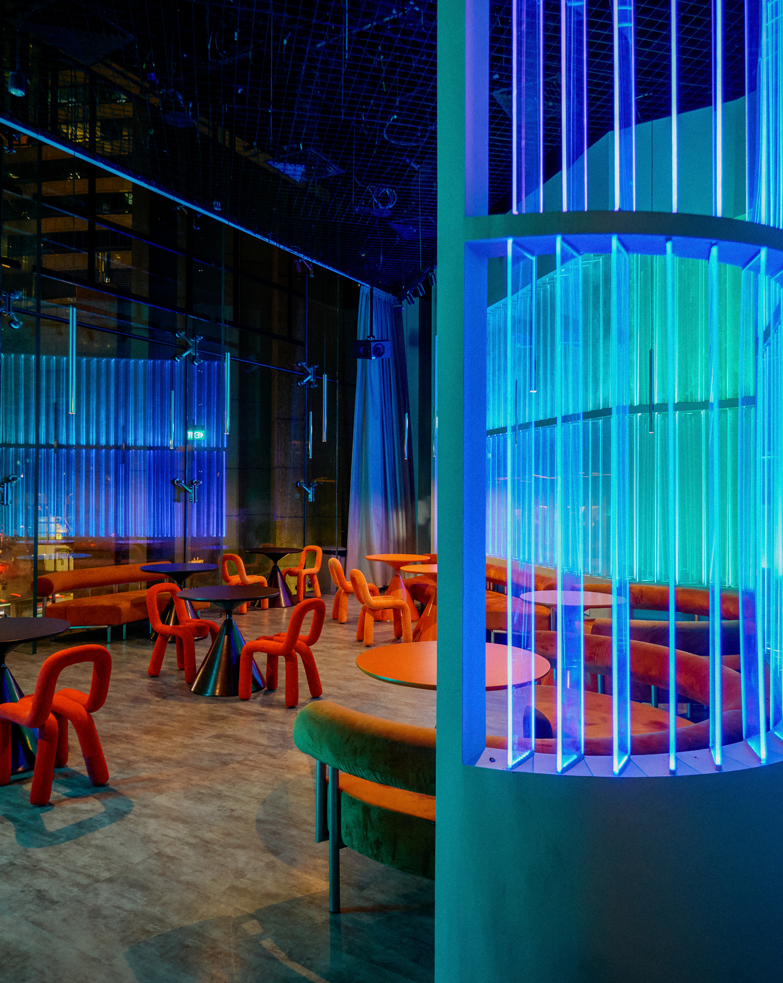 Interior of a modern lounge or bar with blue neon lighting, orange chairs, and black tables, featuring a curved illuminated blue and green installation.