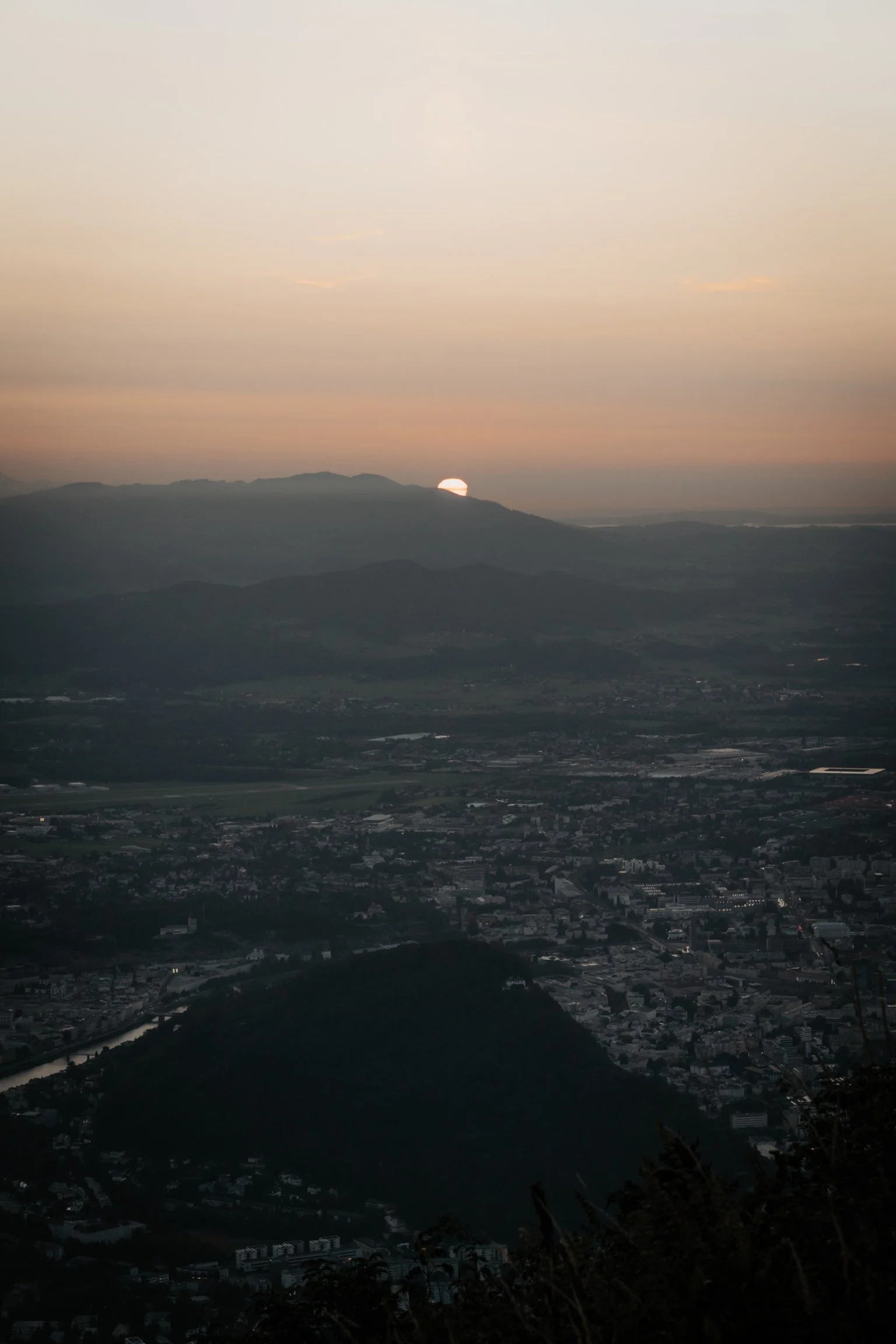 Blick auf eine Sonnenuntergangsszene über Bergen und eine Stadt, die in Dämmerung gehüllt ist.