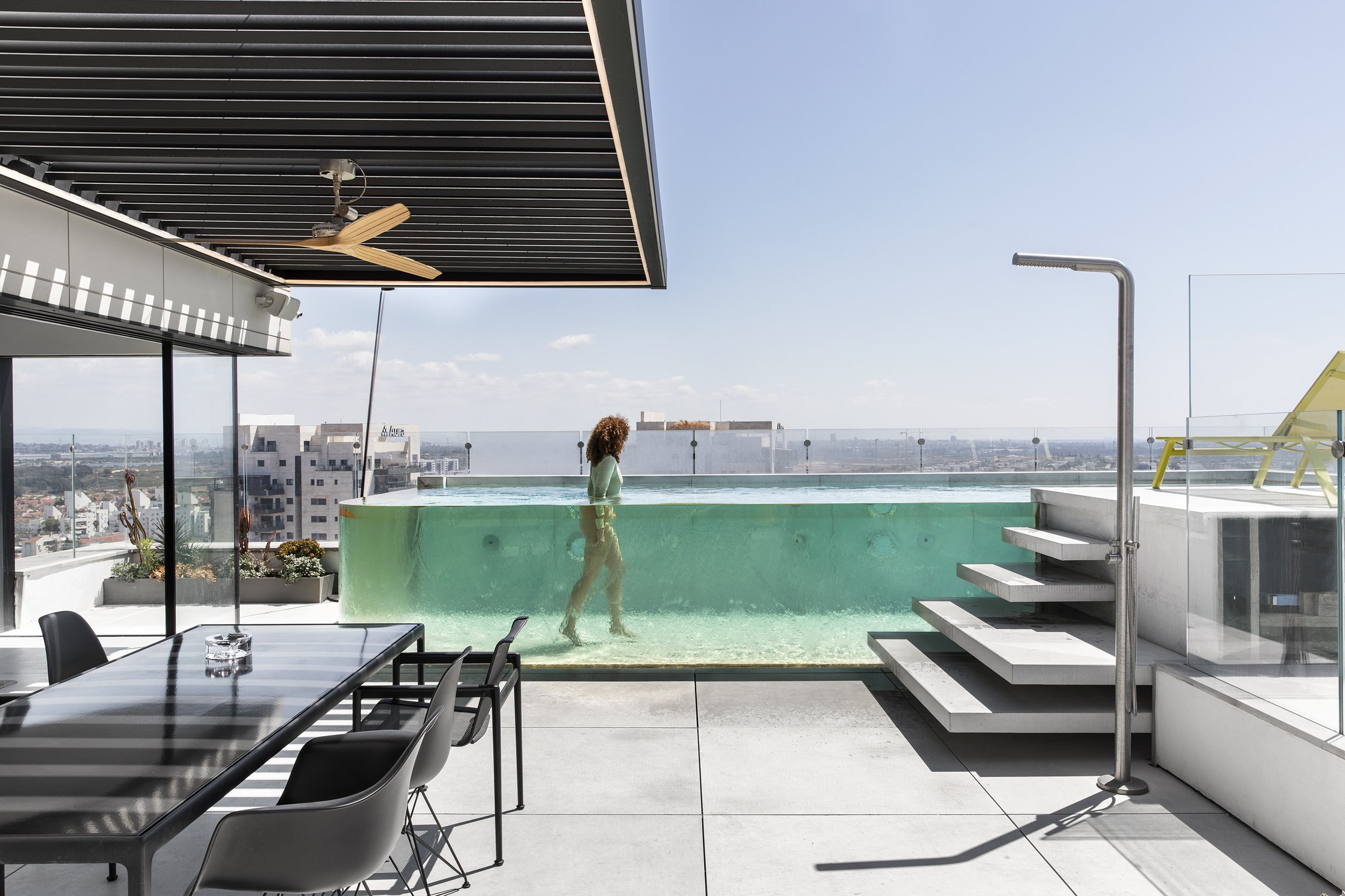 Person in an outdoor rooftop pool on a sunny day with city skyline views, glass railing, modern furniture, and a shaded area with a ceiling fan.