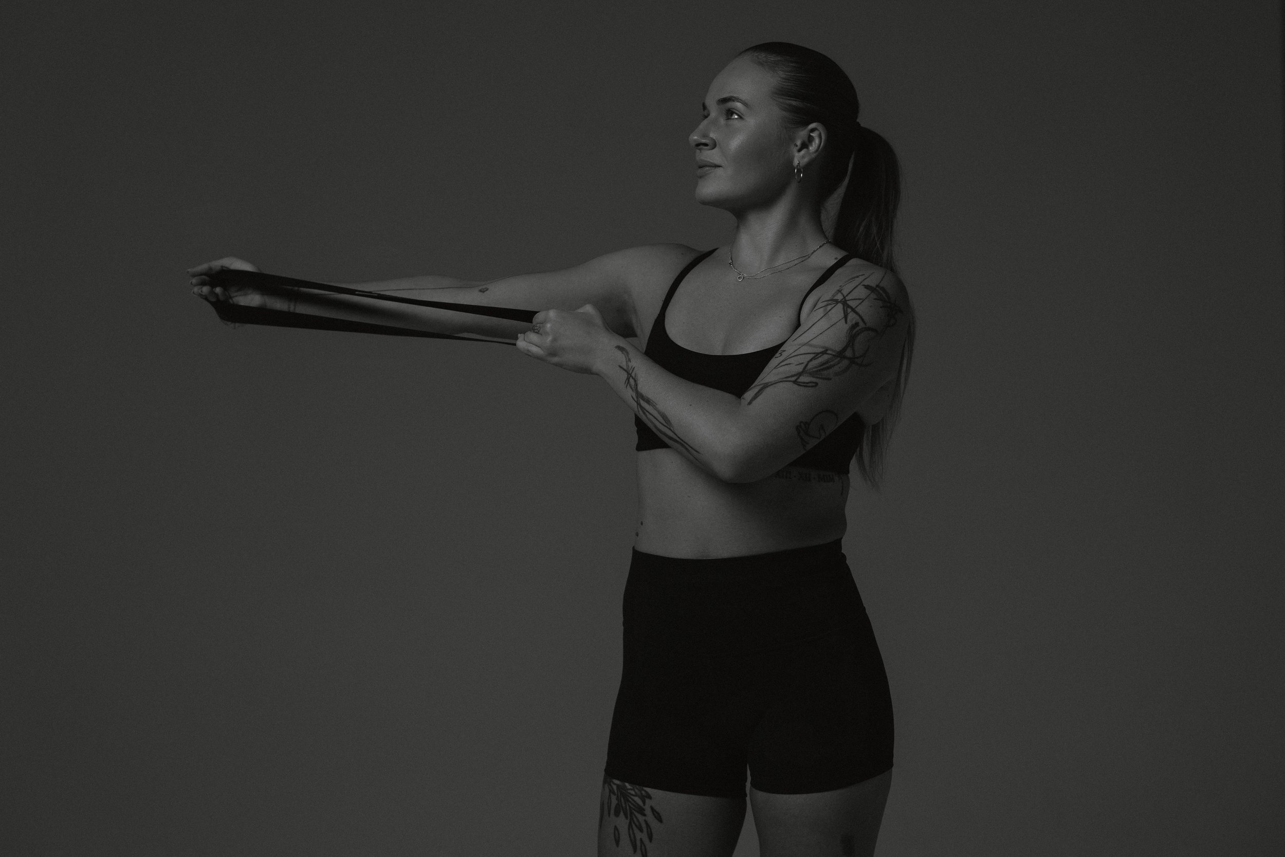 A woman with tattoos on her arms and leg extending her arms forward holding resistance bands, wearing a sports bra and shorts, against a plain background.