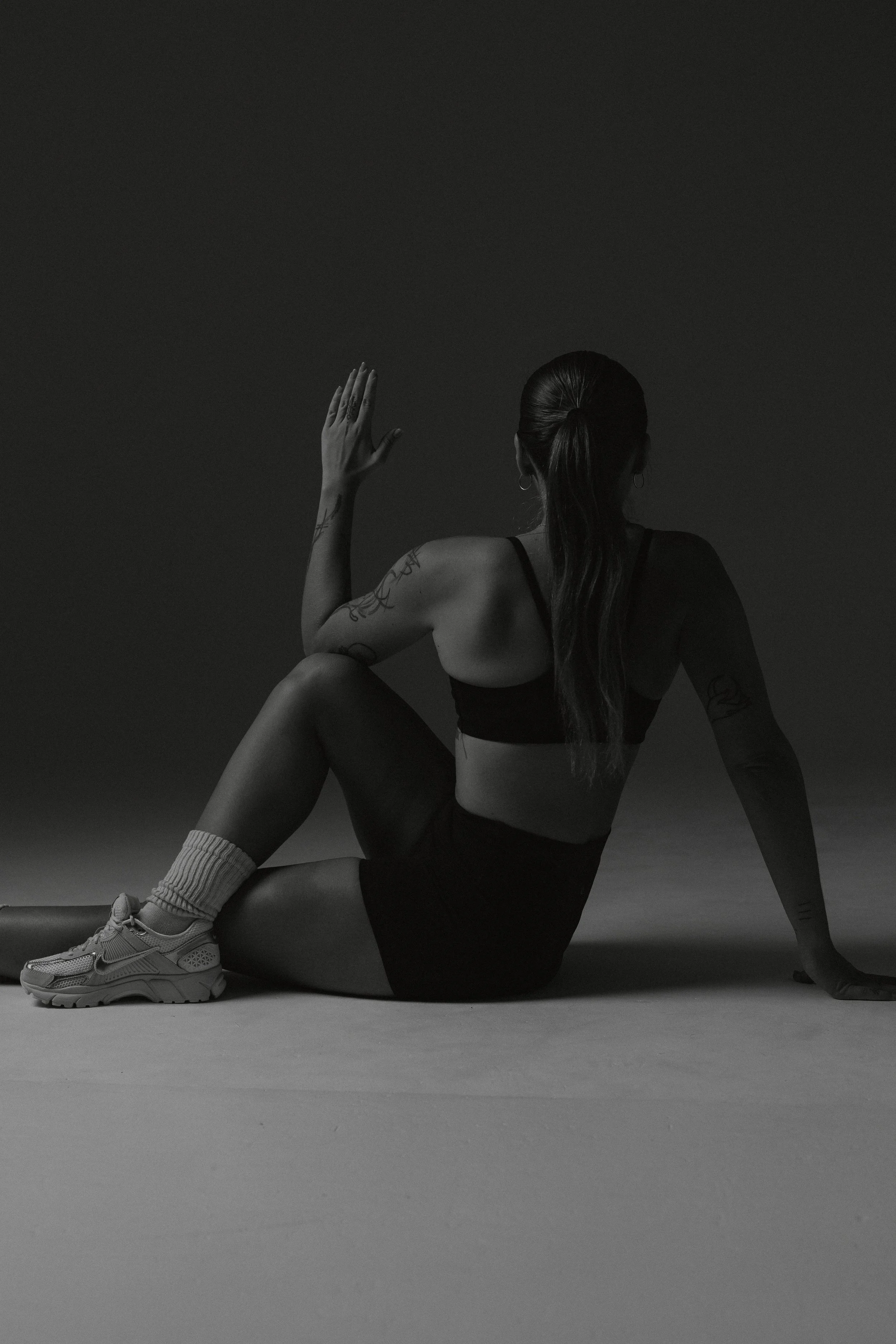 Black and white photo of a woman sitting on the floor with her back turned, wearing a sports bra, shorts, and sneakers, against a dark background.
