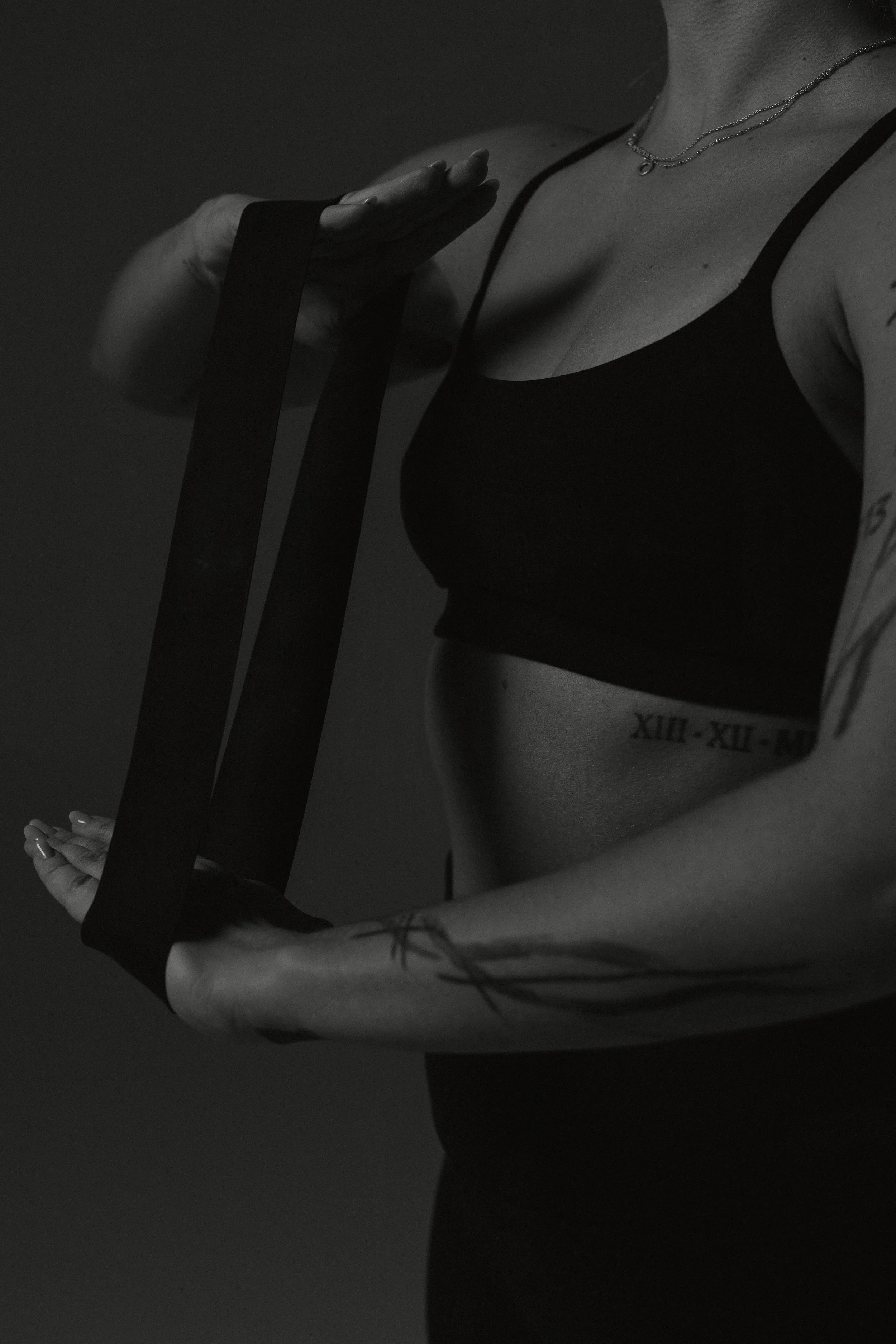 A woman in a sports bra holds a resistance band across her chest, with tattoos on her arm and a tattooed Roman numeral tattoo on her side.