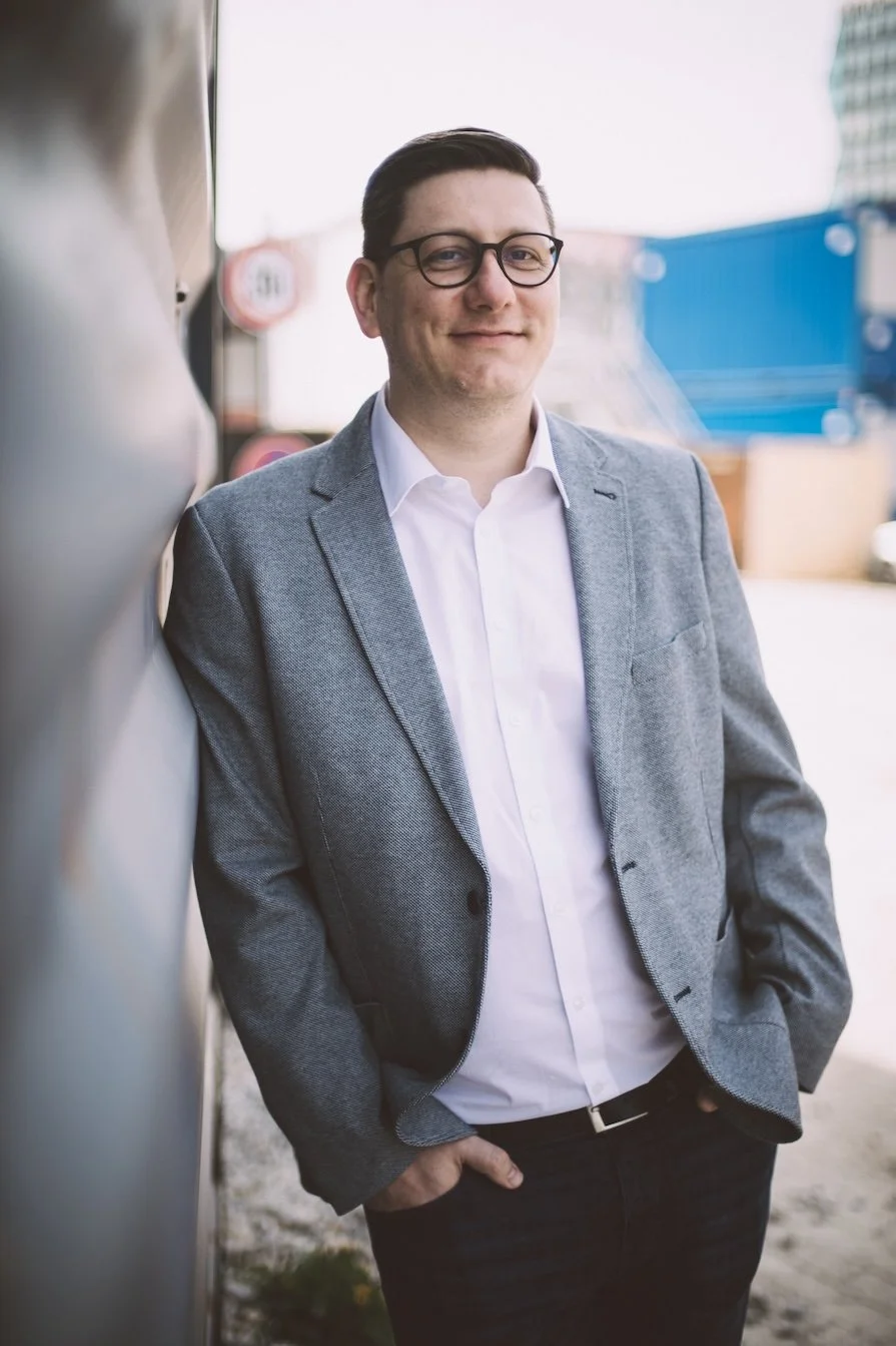 Ein Mann in formeller Kleidung, graues Sakko und weiße Hemd, lächelnd draußen vor einem modernen Gebäude.