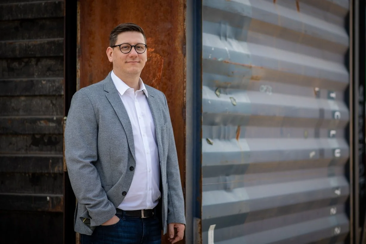 Ein Mann in einem grauen Blazer und weißem Hemd steht vor einer Wand aus Metallpaneelen.