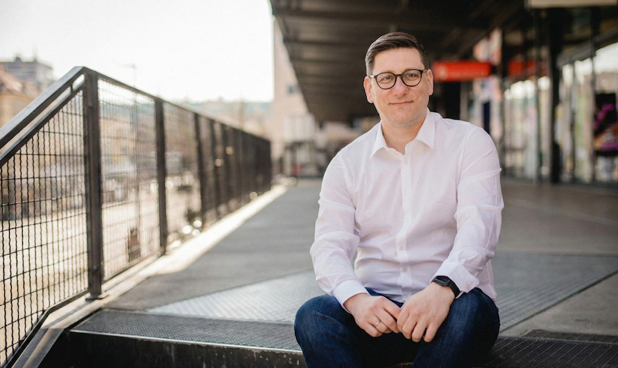 Ein Mann mit Brille, weißem Hemd und dunklen Hosen sitzt draußen auf einer Treppe an einer Stadtpromenade, lächelt in die Kamera.