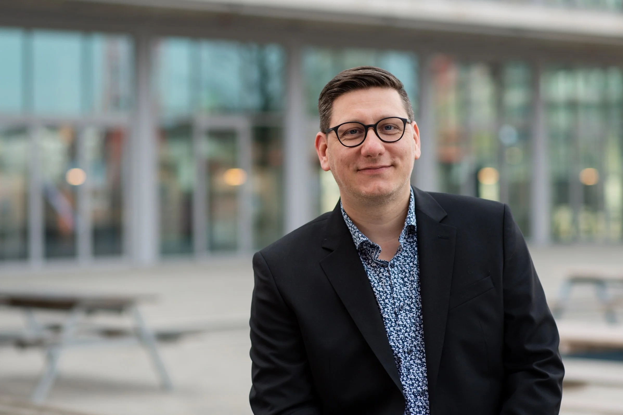 Ein Mann mit Brille, dunklem Anzug und Hemd mit Muster sitzt in einem modernen Gebäude mit viel Glas. Er lächelt leicht.