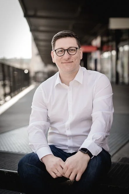 Ein Mann mit Brille, weißem Hemd und dunklen Haaren sitzt draußen auf einer Bank an einer urbanen Straße und lächelt.