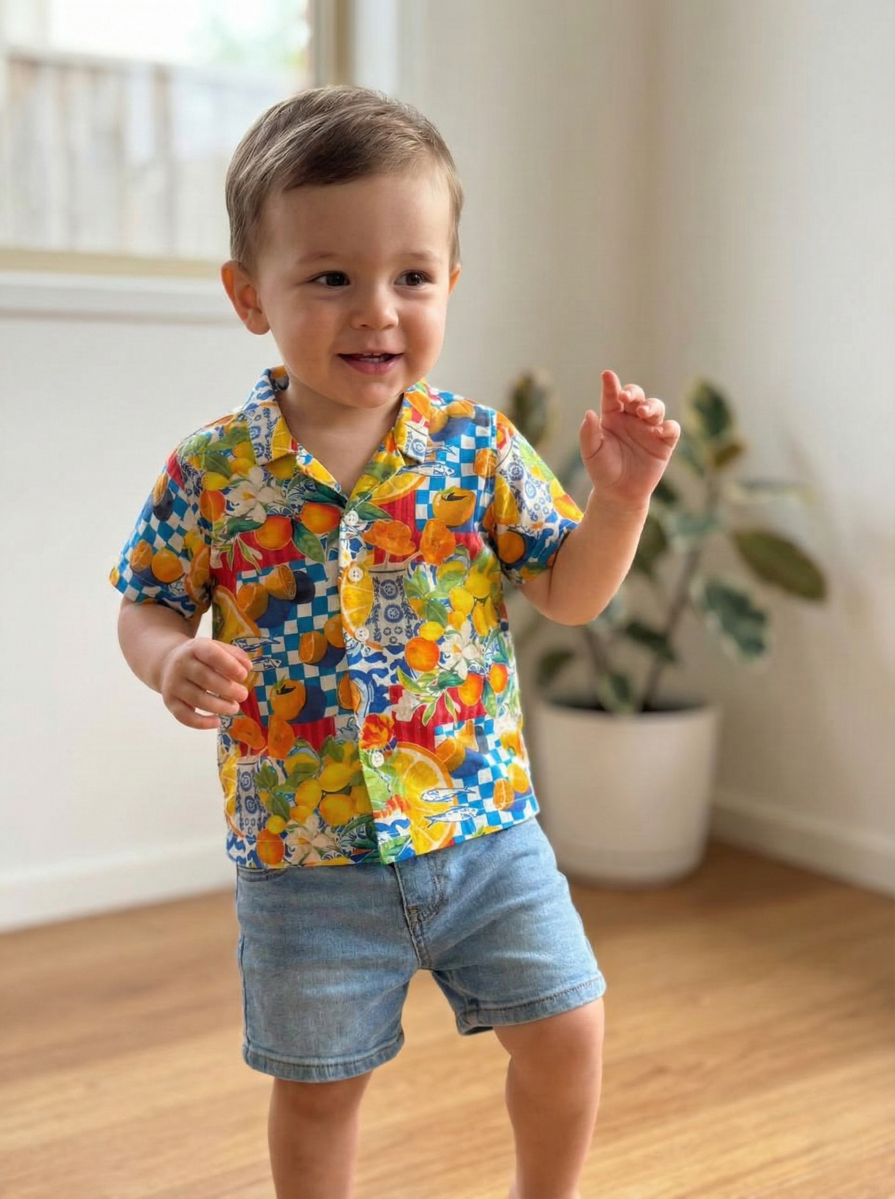 A young boy with brown hair wearing a colorful, short-sleeved Hawaiian shirt and denim shorts, standing indoors near a potted plant.
