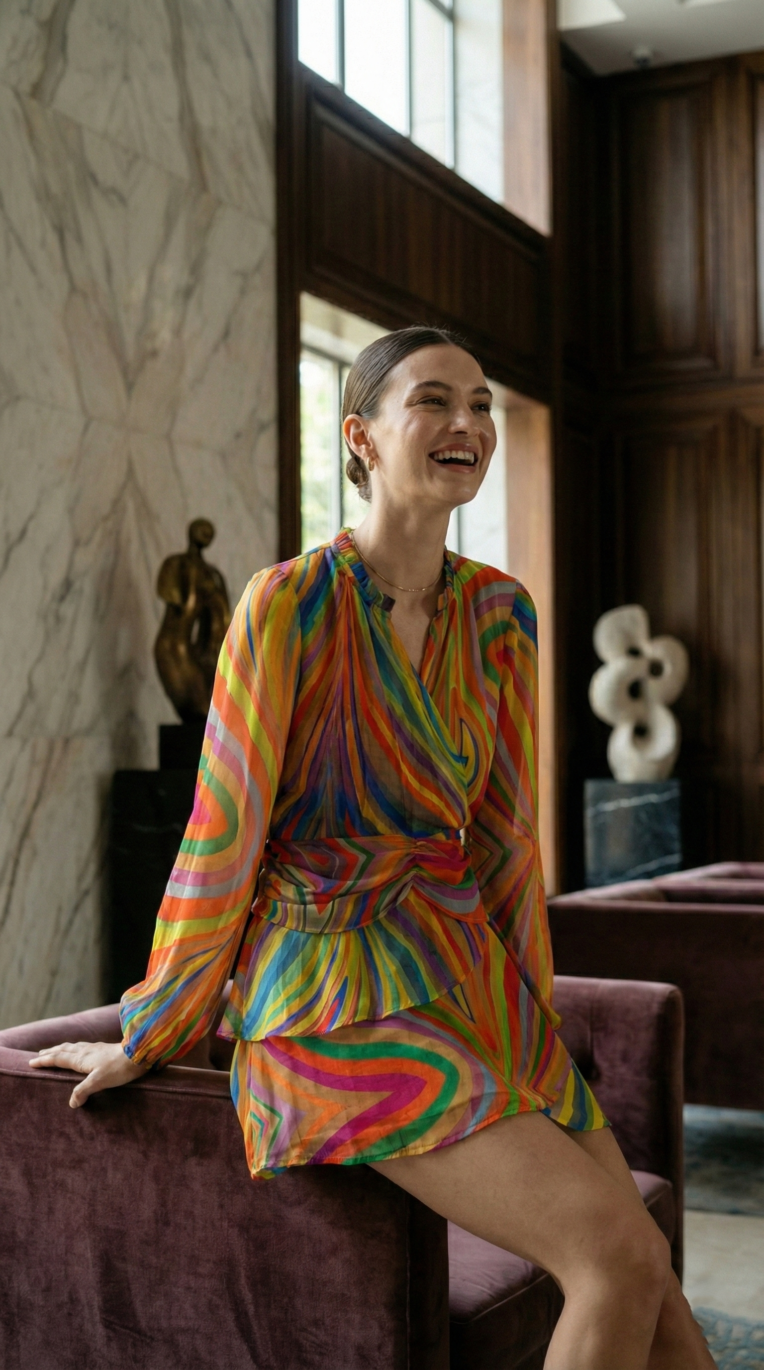 A woman in a colorful, swirling patterned dress sitting on a velvet sofa in a room with art sculptures and large windows.