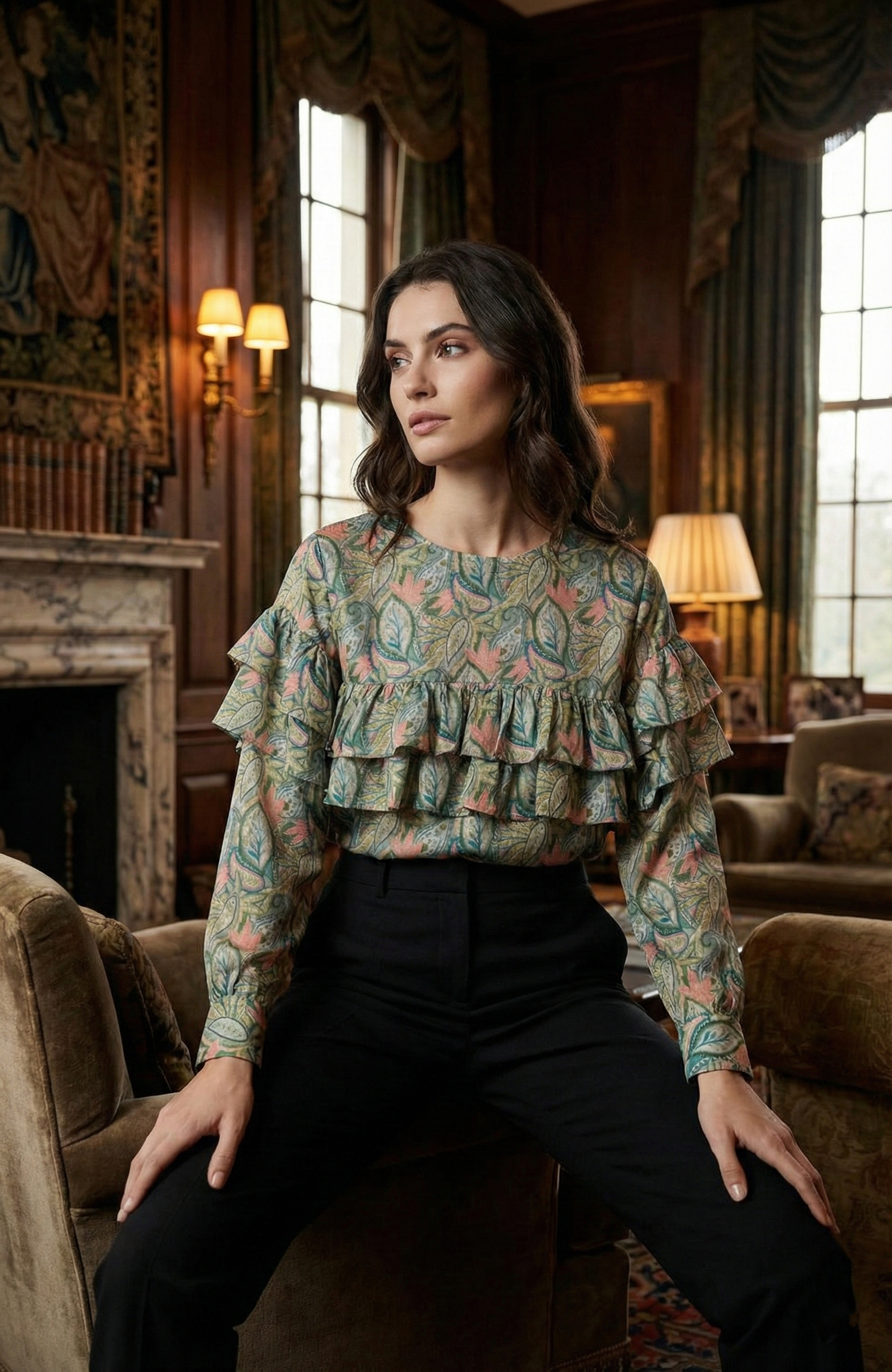 A woman with dark, wavy hair wearing a floral ruffled blouse and black pants sitting in a richly decorated living room with large windows, wooden walls, and warm lighting.