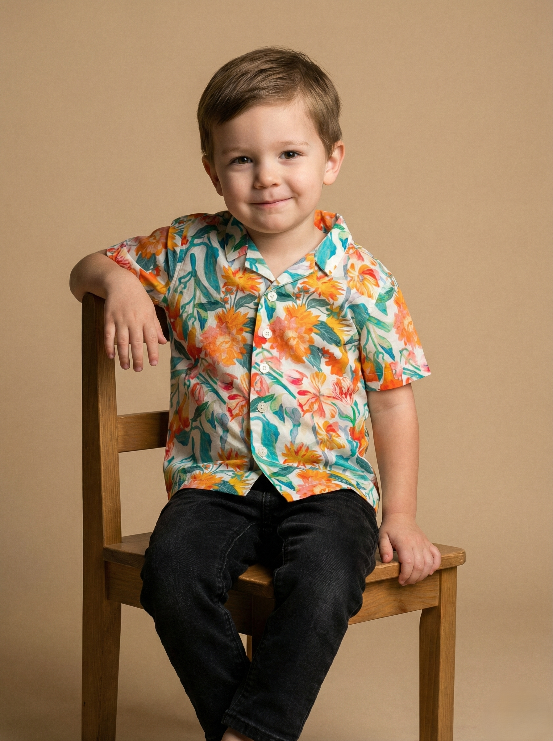 Young boy sitting on a wooden chair, wearing a colorful floral shirt and black pants, smiling gently, in a studio setting with a plain beige background.