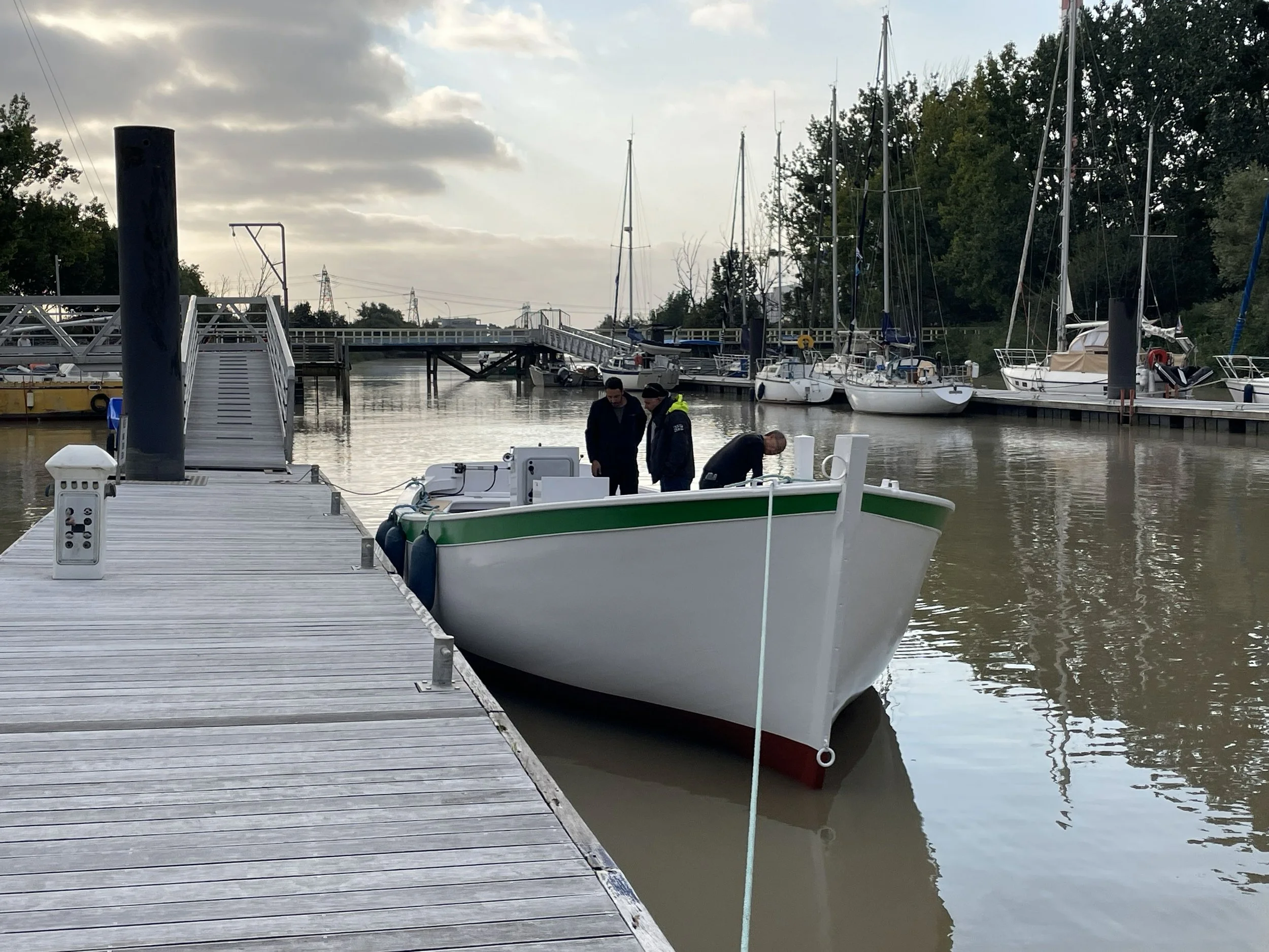 Un ponton en bois avec plusieurs bateaux à voile amarrés, deux personnes en discussion, arbres en arrière-plan et un ciel nuageux.