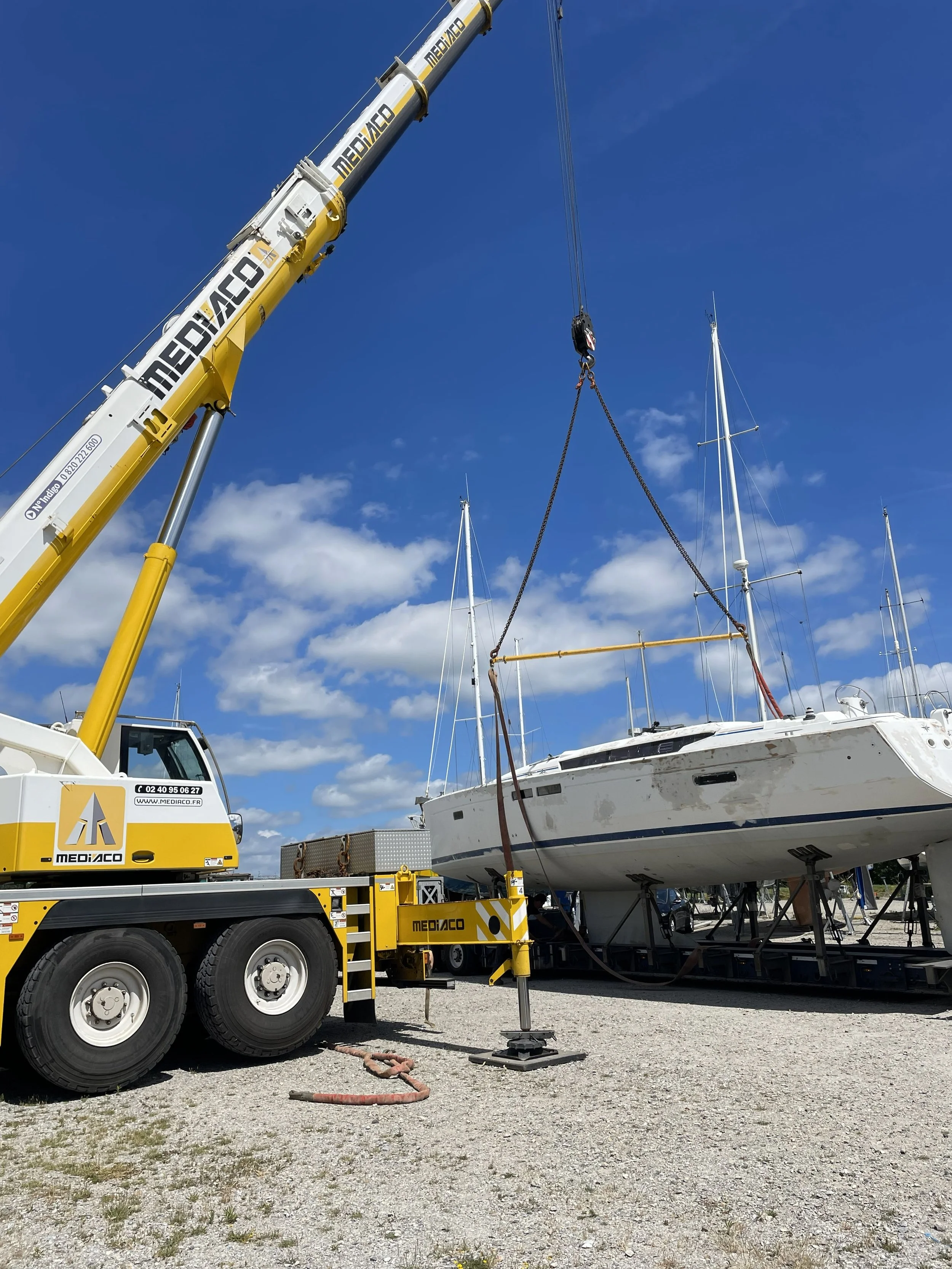 Un bateau est soulevé par un pont roulant jaune sur une surface en gravier, avec un ciel bleu et quelques nuages.