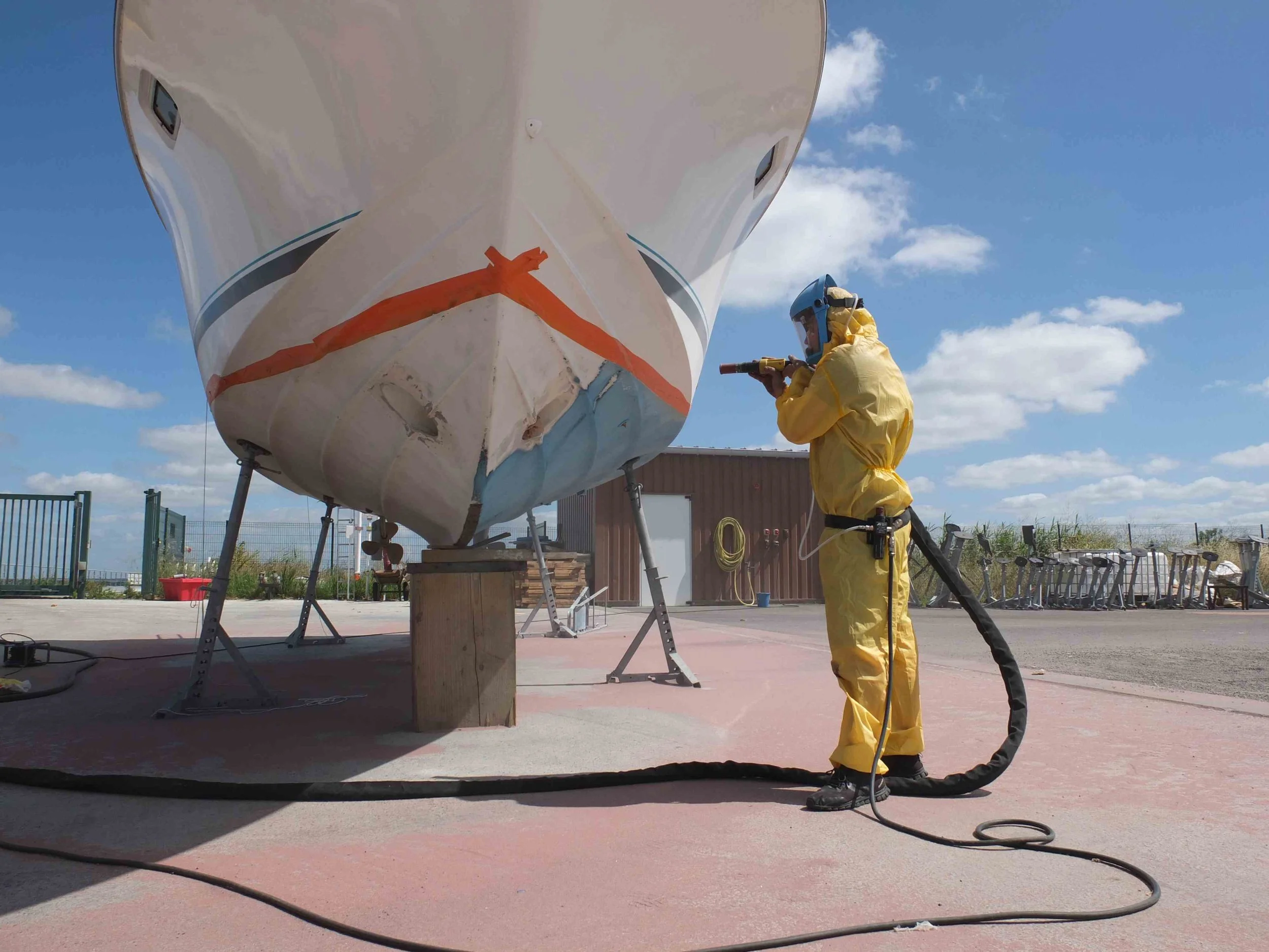 Une personne en combinaison de protection jaune et un casque travaille sur la coque d'un bateau, utilisant un outil électrique, dans une zone d'extérieur.