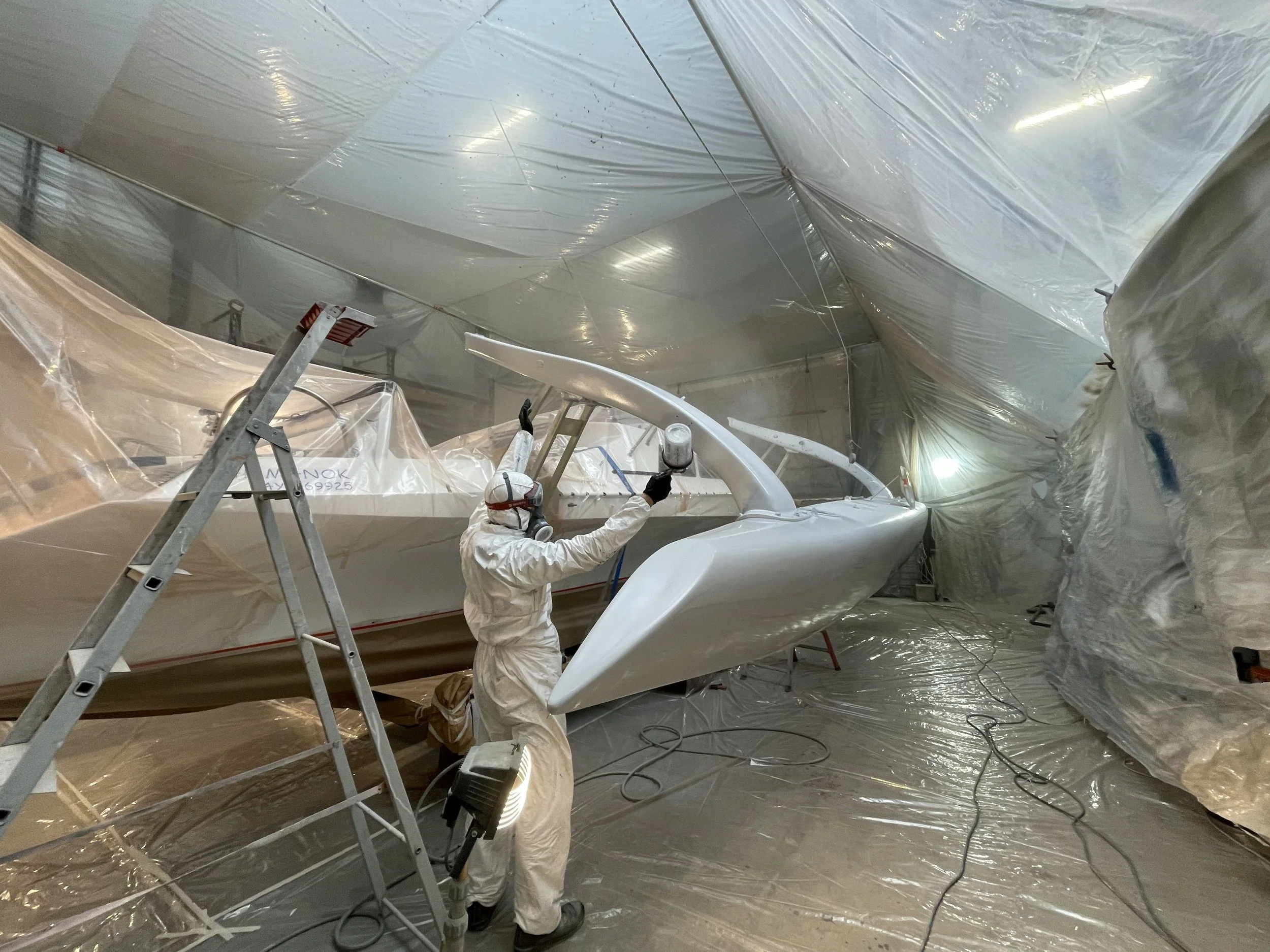 Une personne en combinaison de protection, masquée et portant des gants, travaille sur un bateau en intérieur, entouré de plastique de protection pour la peinture ou la réparation.