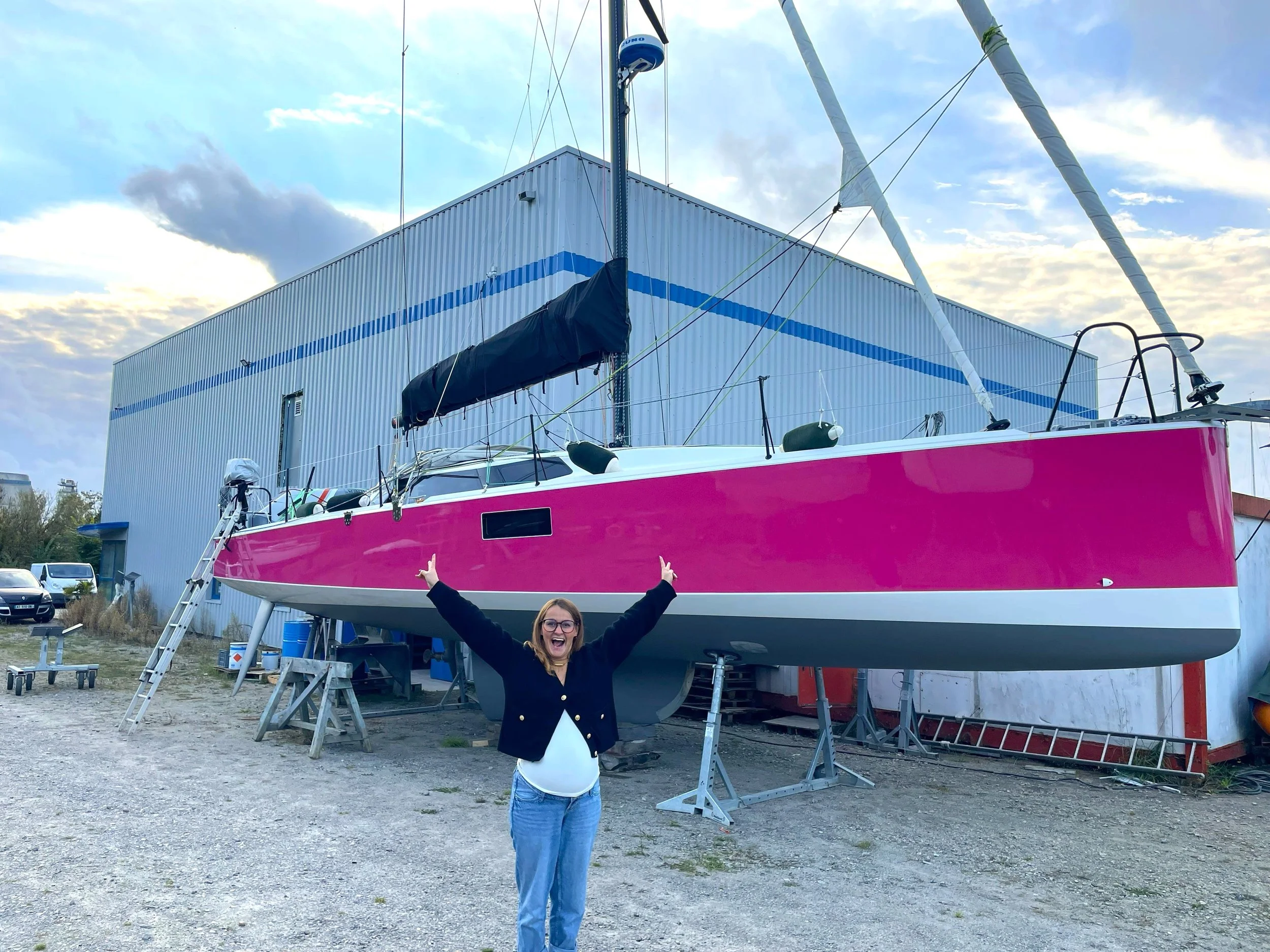 Une femme souriante avec des lunettes, levant les bras, se tient devant un voilier large de couleur rose, en dehors d'un bâtiment industriel, avec plusieurs voitures en arrière-plan.
