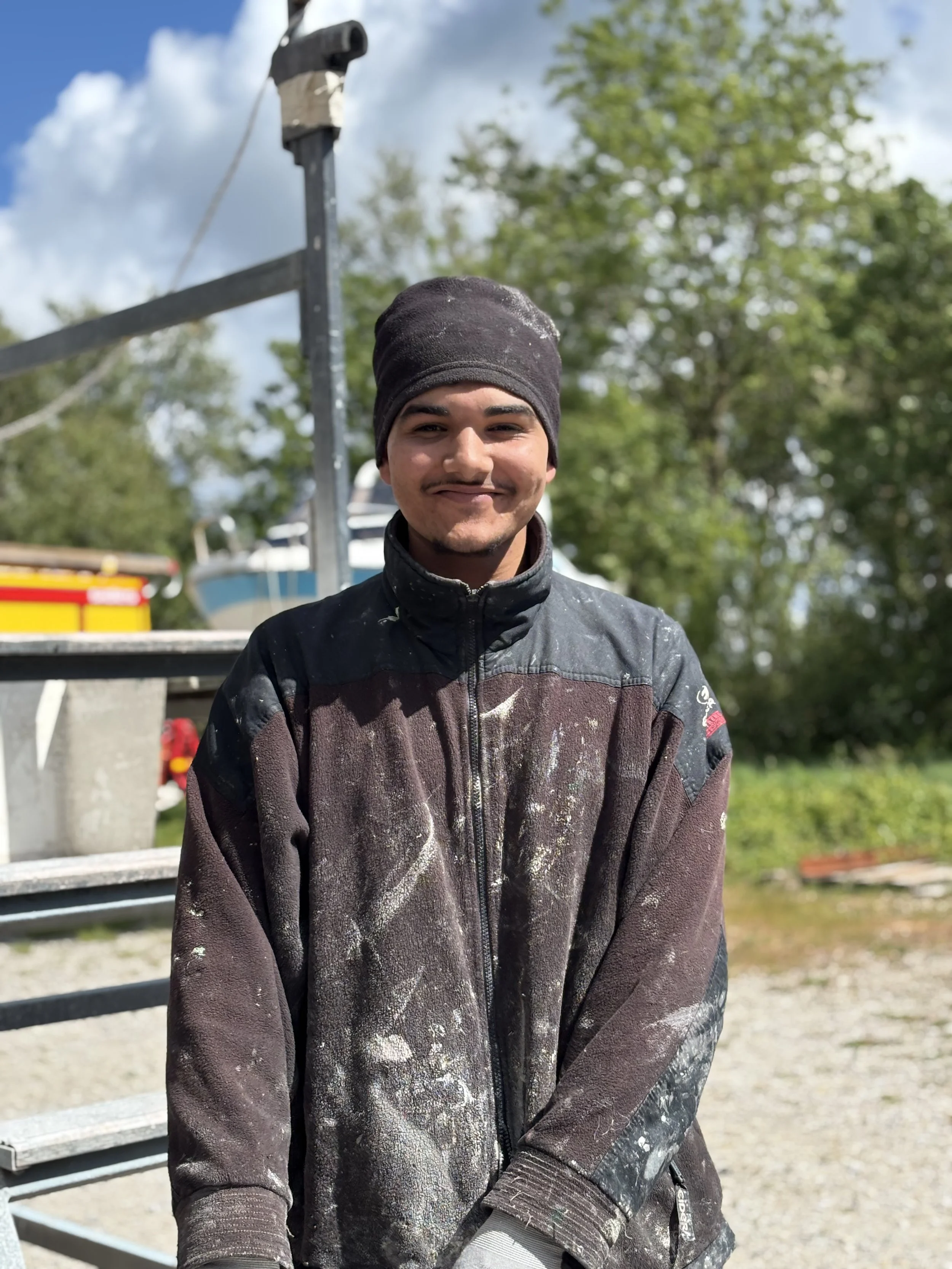 Jeune homme souriant portant une couche de peinture, casquette noire, veste de travail tachée de peinture, debout à l'extérieur avec un ciel bleu et des arbres en arrière-plan.