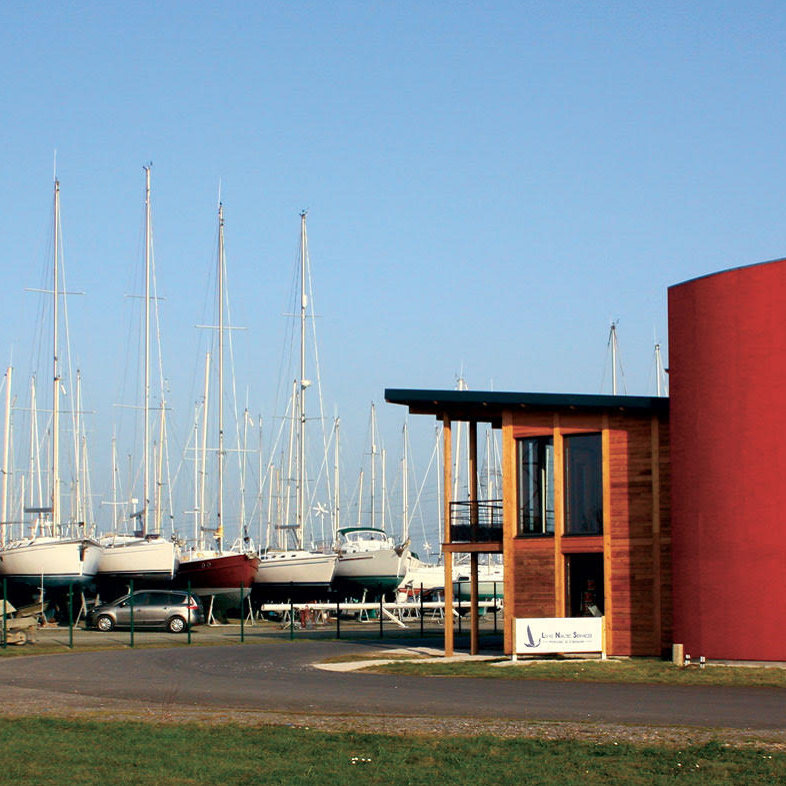 Vue d'un port avec plusieurs voiliers amarrés et un bâtiment en bois moderne à côté, sous un ciel clair.