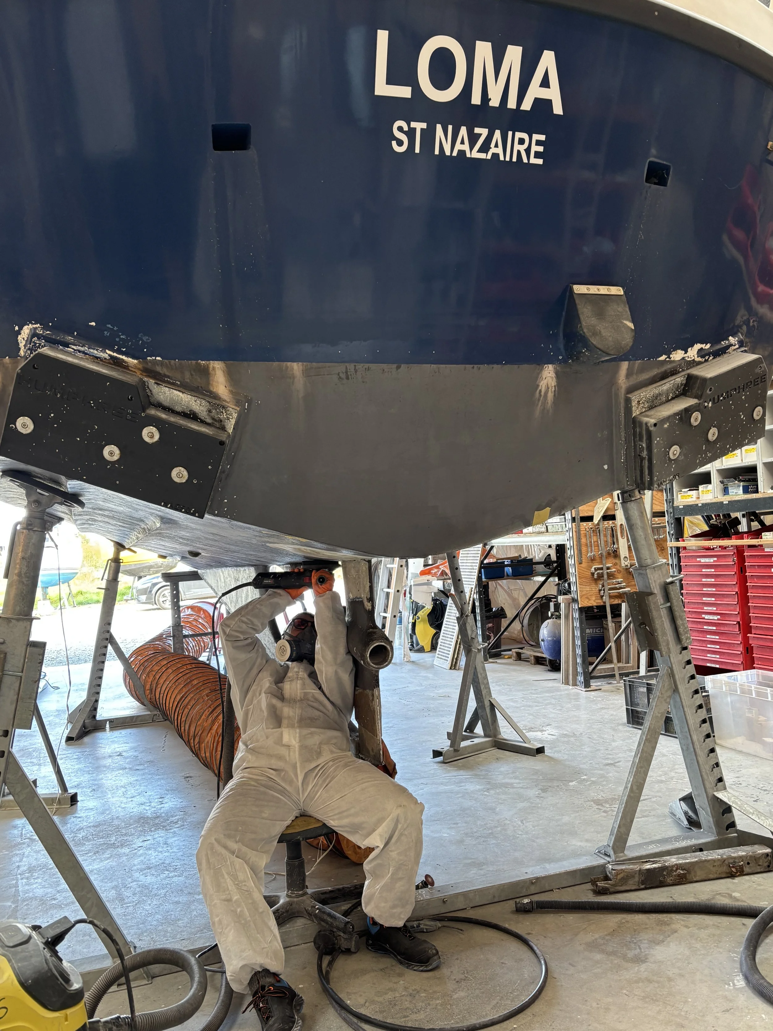 Une personne en tenue de protection travaillant sur la coque d'un bateau, dans un atelier ou un hangar, avec divers outils et équipements autour.