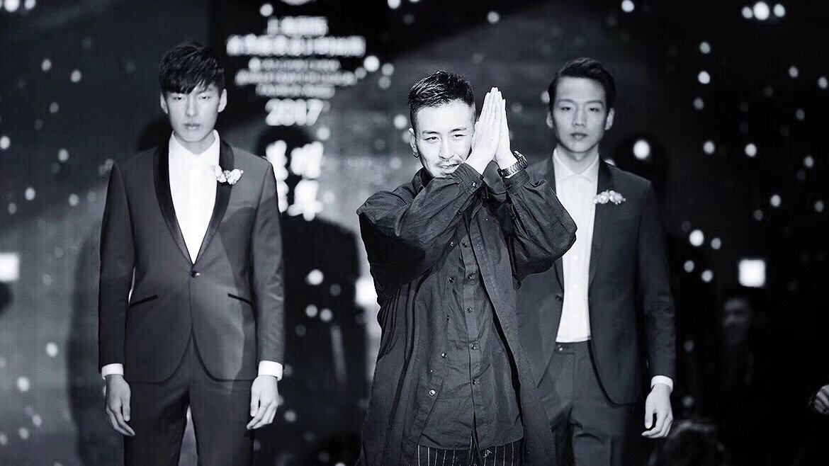 Three men on stage, two in suits, and one in a casual dark jacket, with the man in the middle making a praying gesture with his hands, against a dark, starry background.