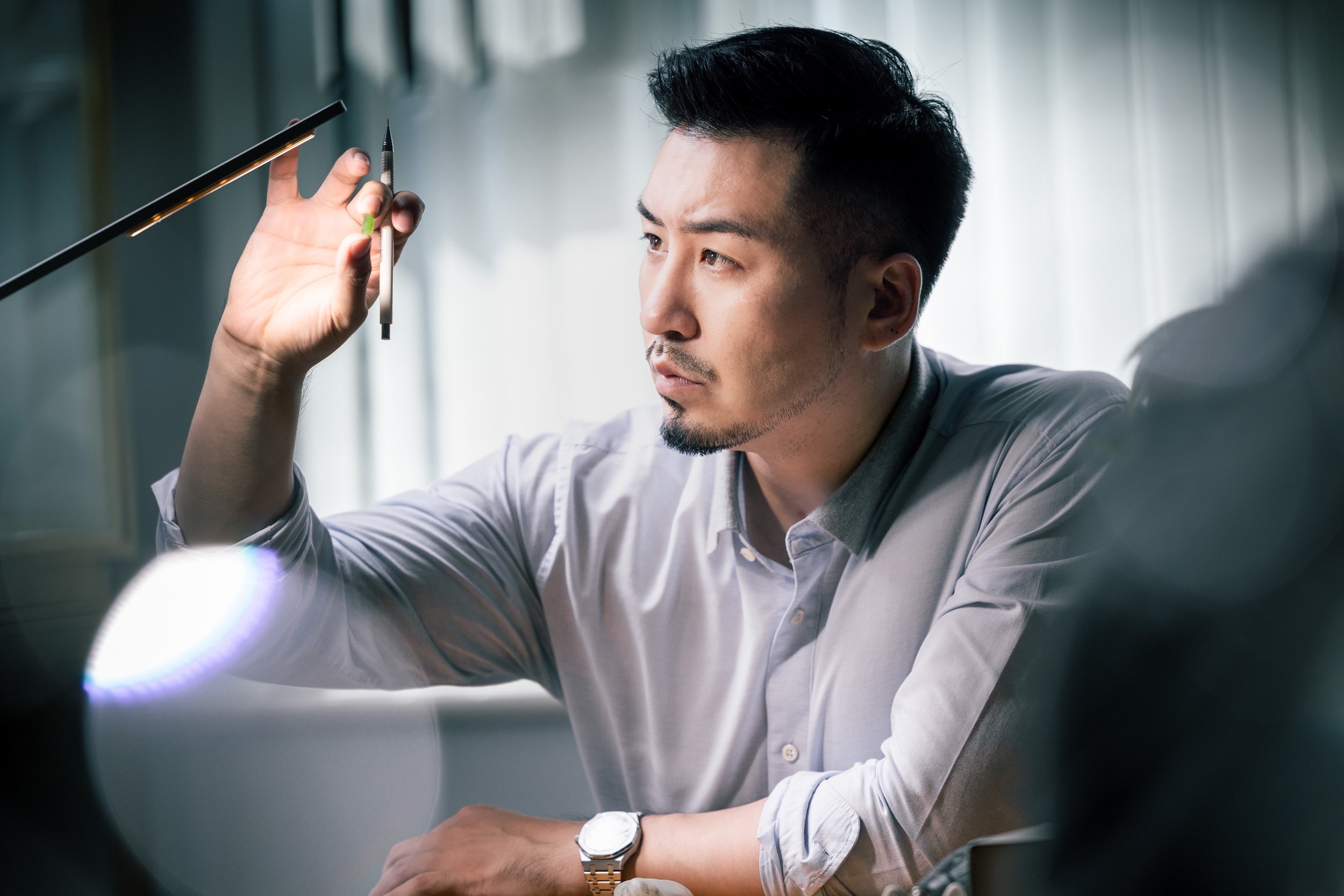 A man with dark hair and a gray shirt sits at a table, holding a pen and examining it closely with a focused expression in a well-lit room.