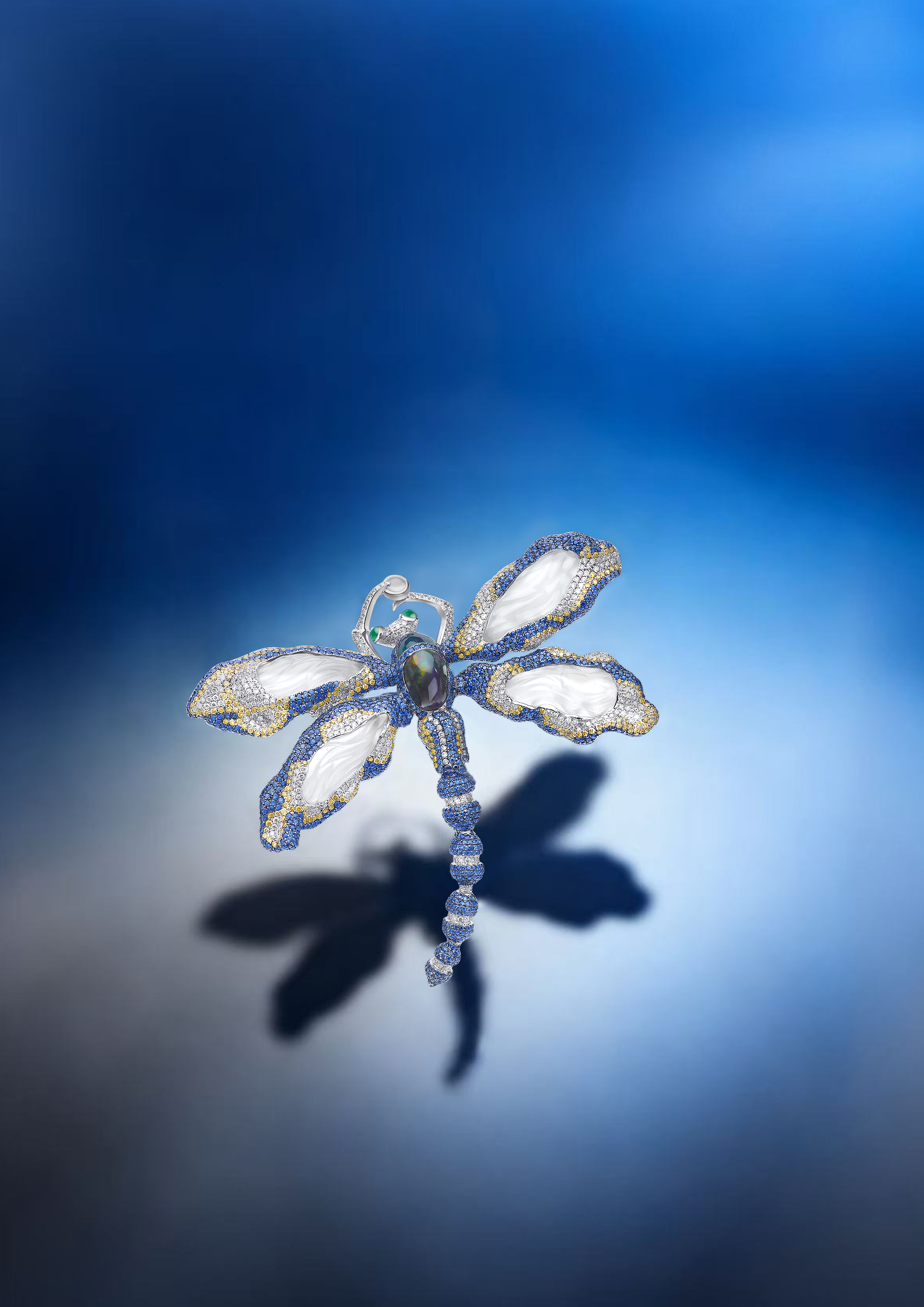 A decorative dragonfly-shaped jewelry piece with blue and gold accents, white wings, and an iridescent body, casting a shadow on a blue gradient background.