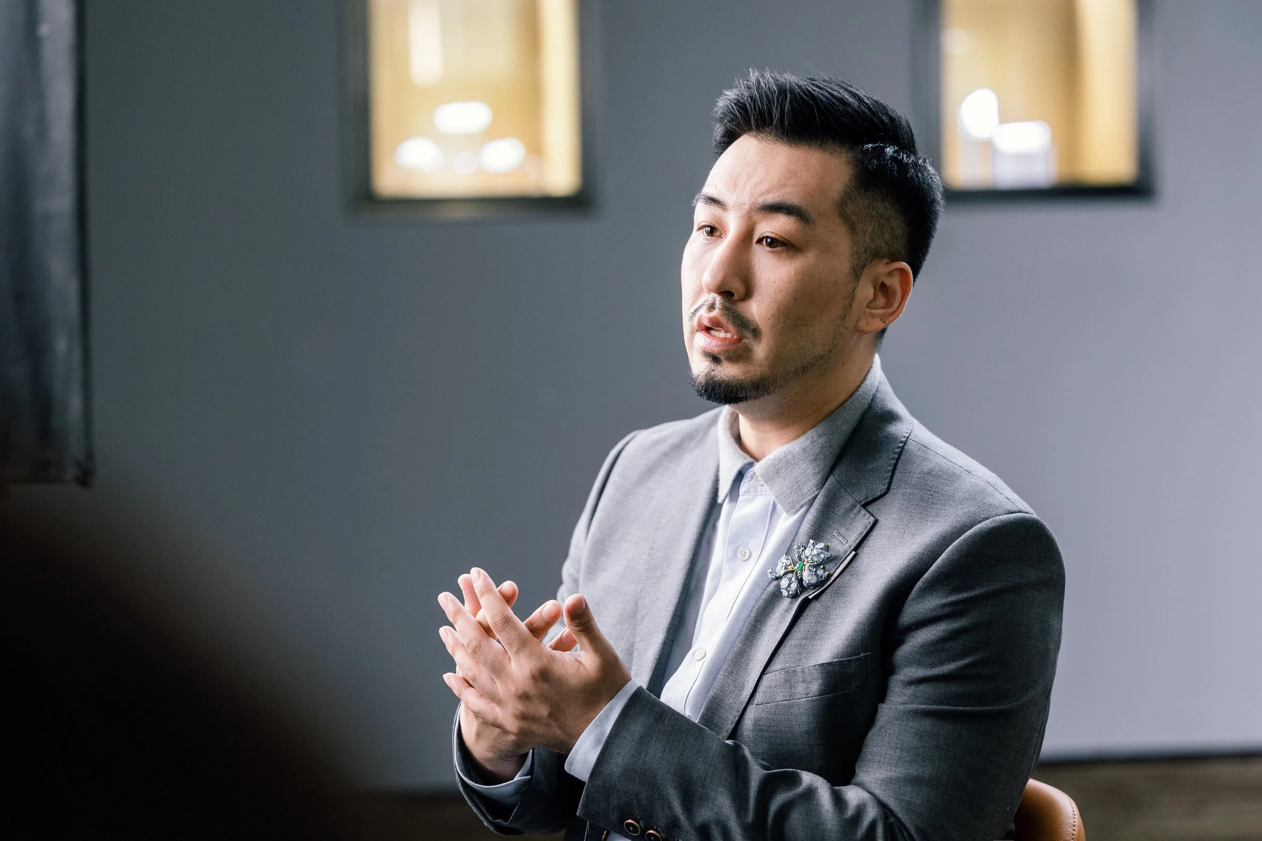A man with black short hair and a beard, wearing a grey suit jacket with a brooch pinned to his chest, sits in a grey-walled room while being interviewed.