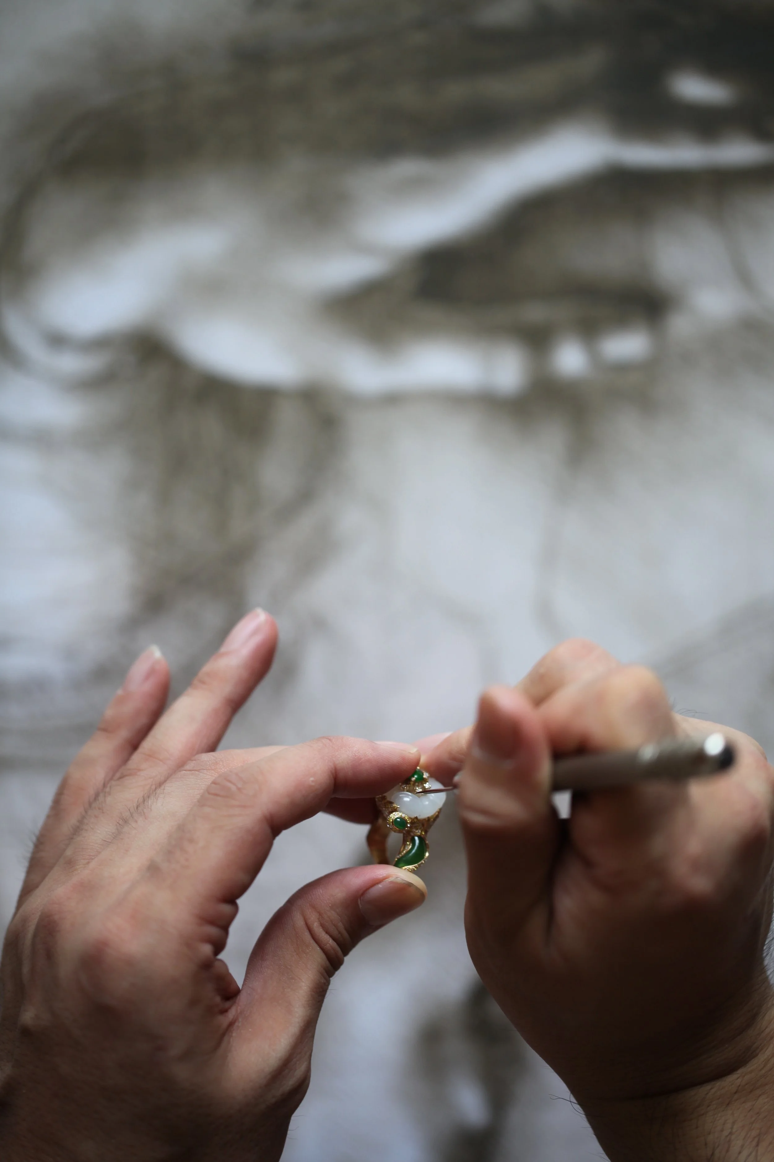 A person is working on jewelry, holding a ring with a pin or small tool, against a blurred background that appears to be a reflective surface or water.