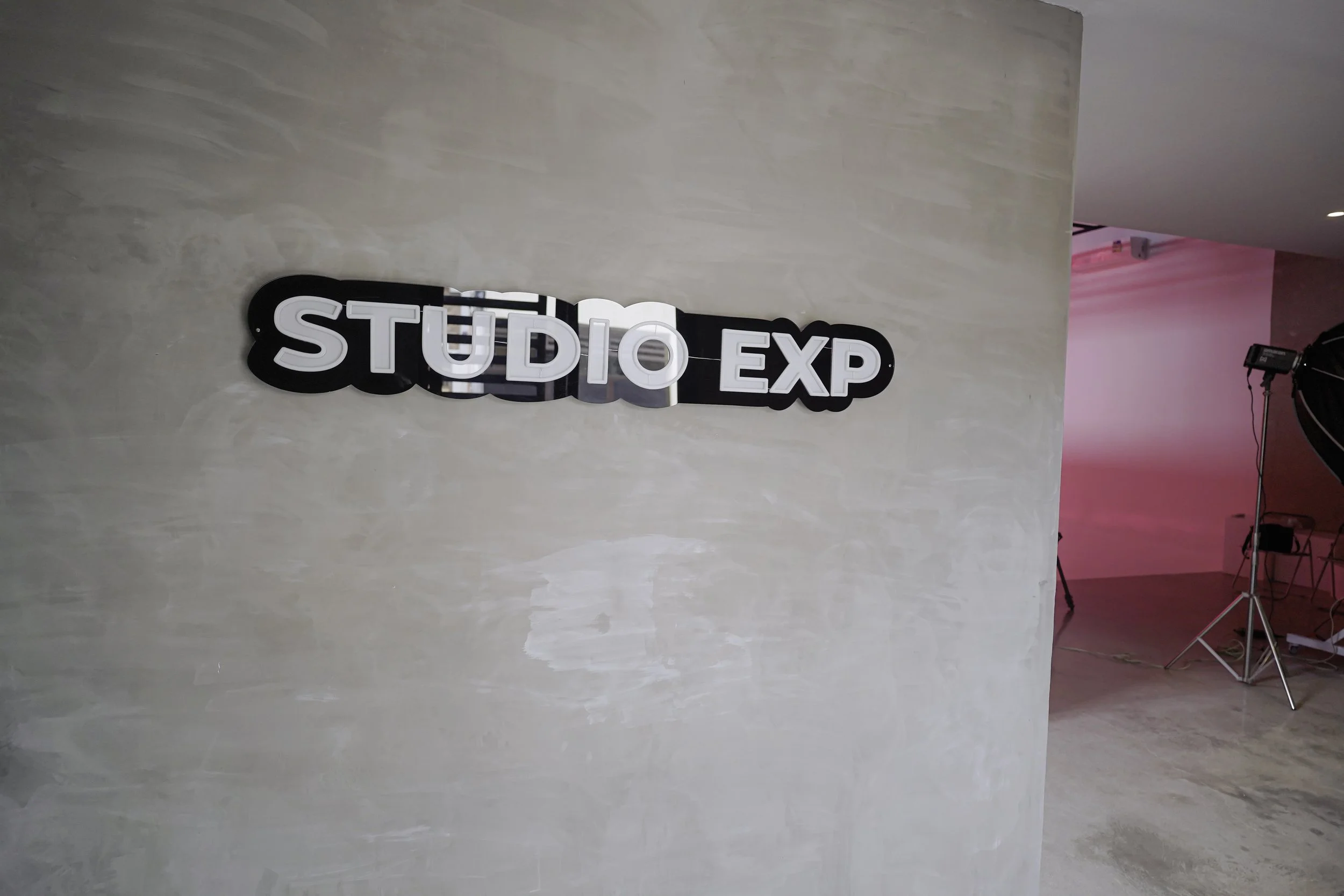 A black and white illuminated sign reading 'STUDIO EXP' on a beige textured wall, with a pink-lit studio space with professional lighting and equipment in the background.