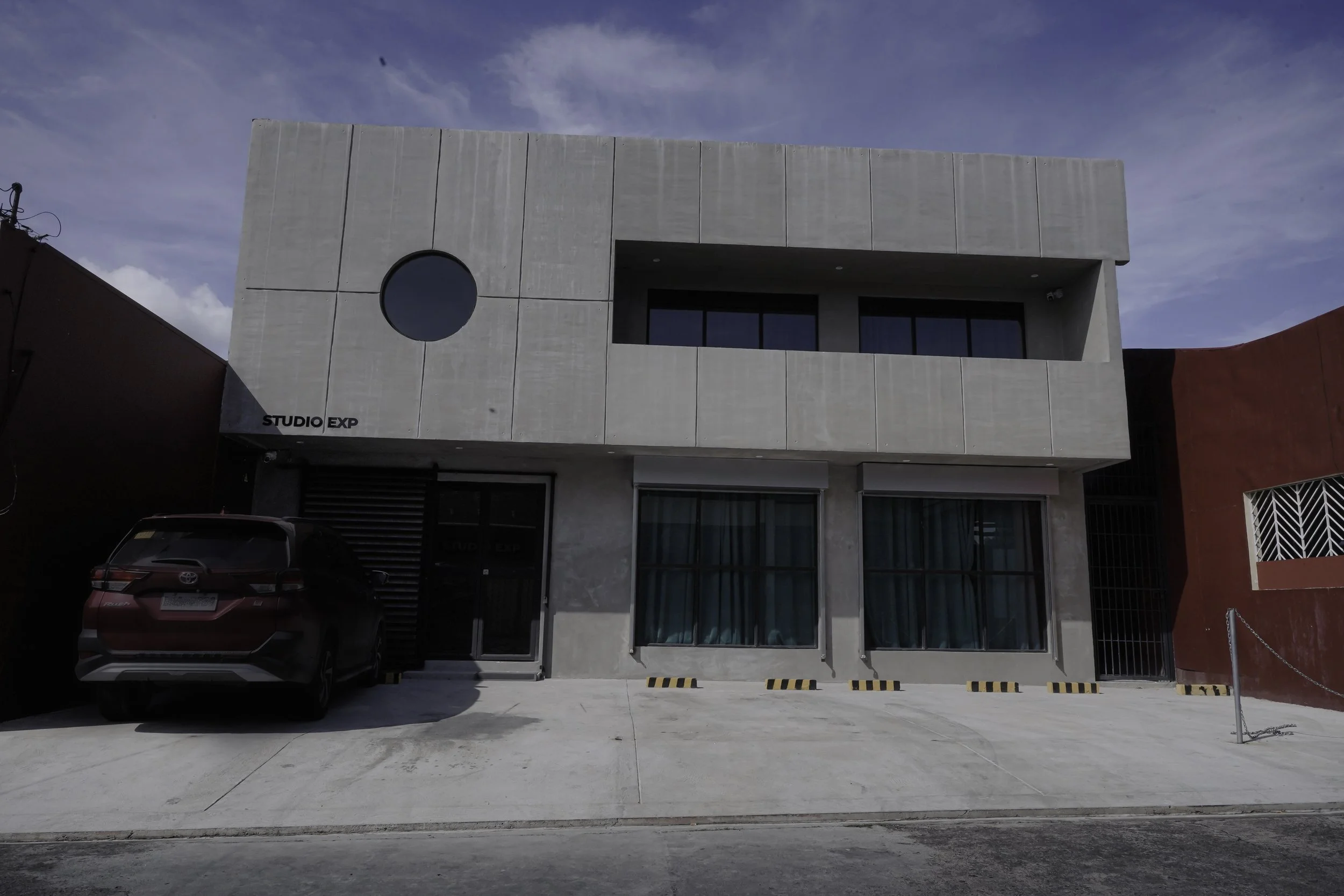 A modern two-story building with a concrete facade, large glass windows, and a circular window near the left corner. There is a black car parked in front and the building is labeled 'STUDIO EXP' on the left side.