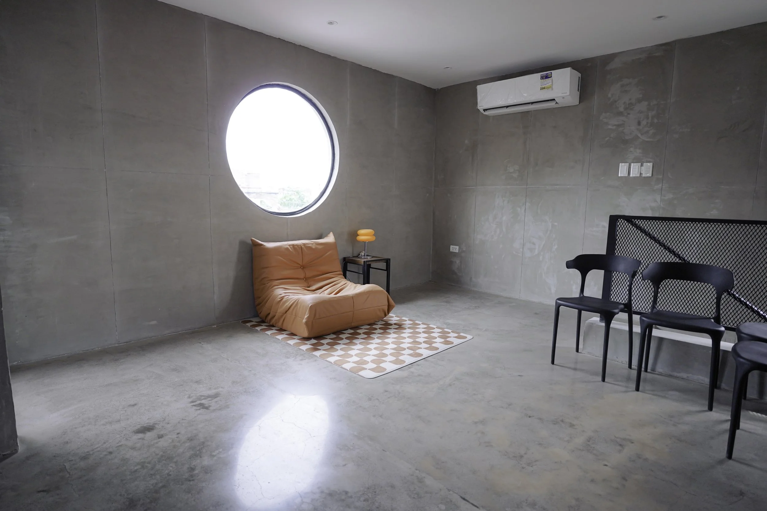 Minimalist living space with a pink low chair on a patterned rug, small side table with an orange lamp, black chairs, concrete walls, a large round window, and an air conditioning unit.