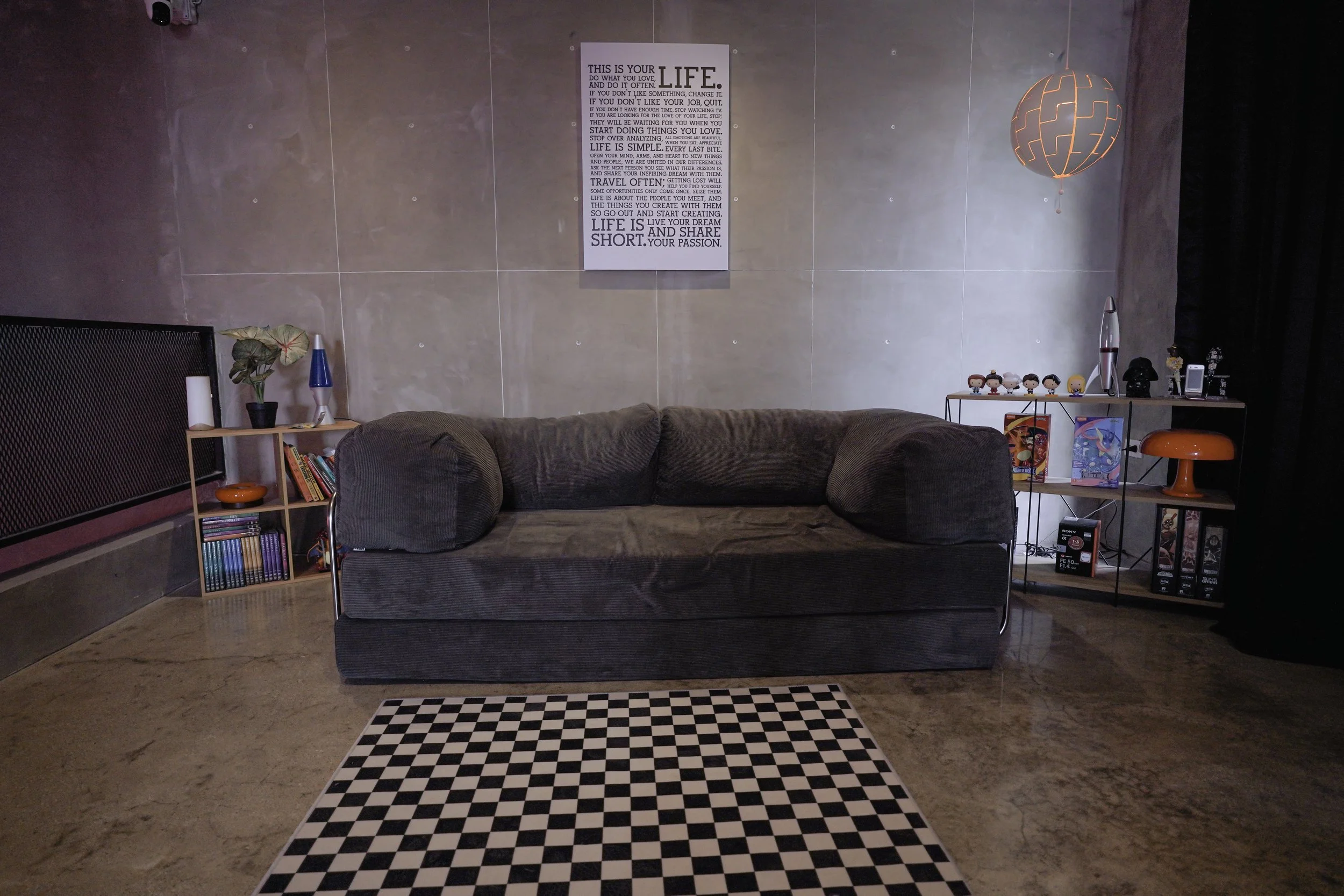 A living room with a dark gray sofa, a checkered black and white rug in front, and two set of shelves with books, figurines, and decor. A wall art with a motivational quote is centered above the sofa, and an orange globe-shaped pendant light hangs fr