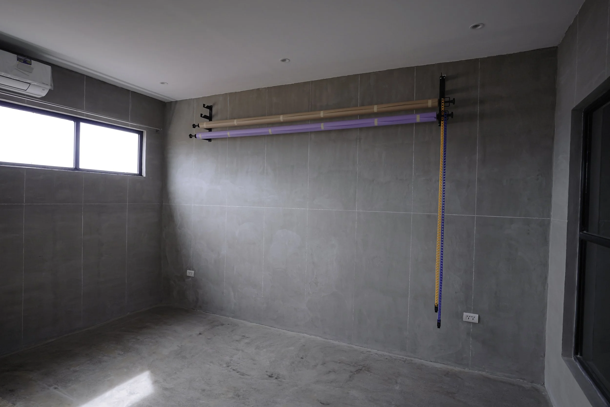 An unfinished room with gray tiled walls and floor, a window on the left, an air conditioning unit, and exercise or rehabilitation equipment mounted on the wall including parallel bars and a measuring tape hanging from one of the bars.