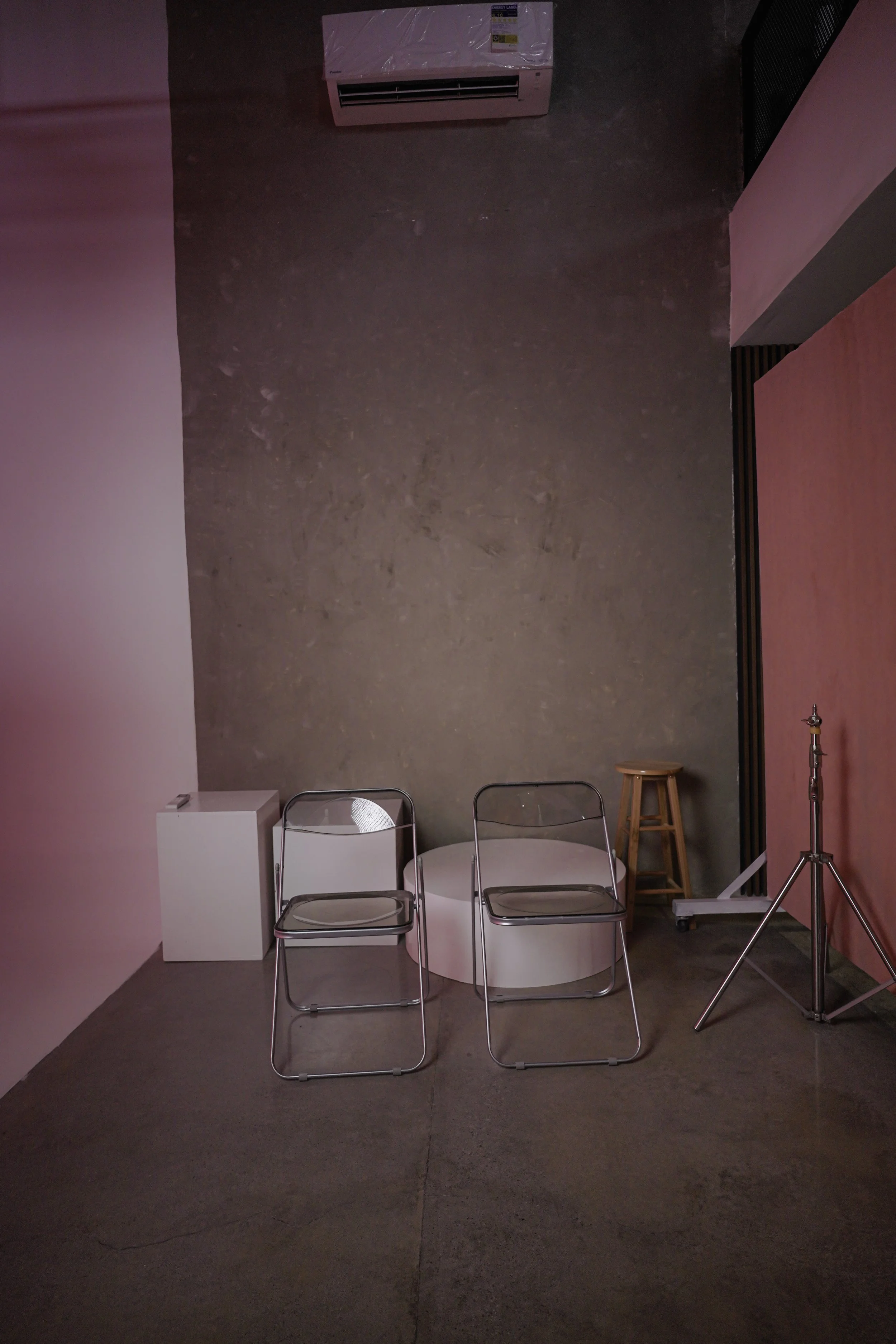Empty room with two metal chairs, a white podium, a wooden stool, and photography equipment against a brick wall with pink lighting