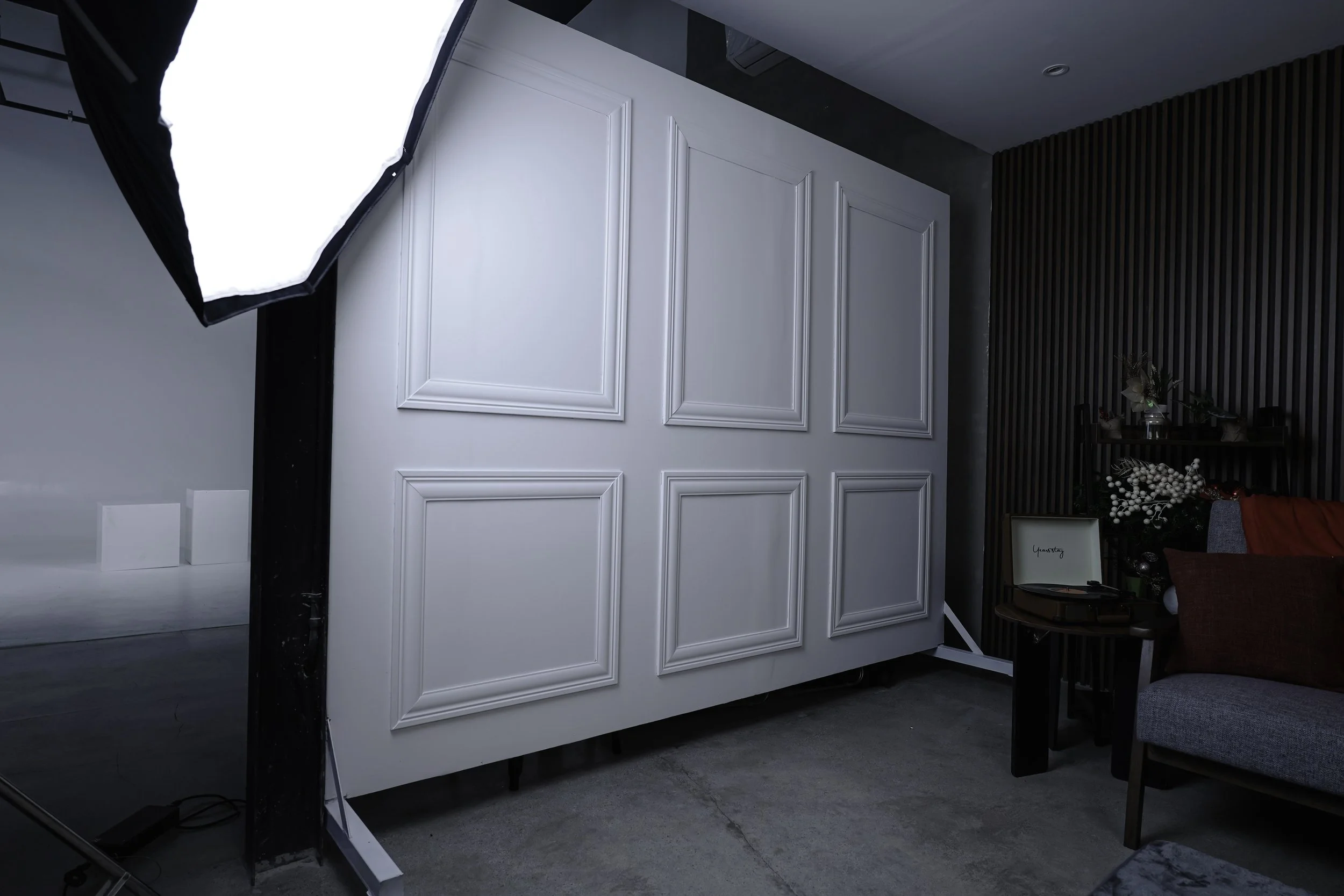 Photography studio setup with a large white backdrop, professional lighting, and a seating area with a small table and decorative items in the background.
