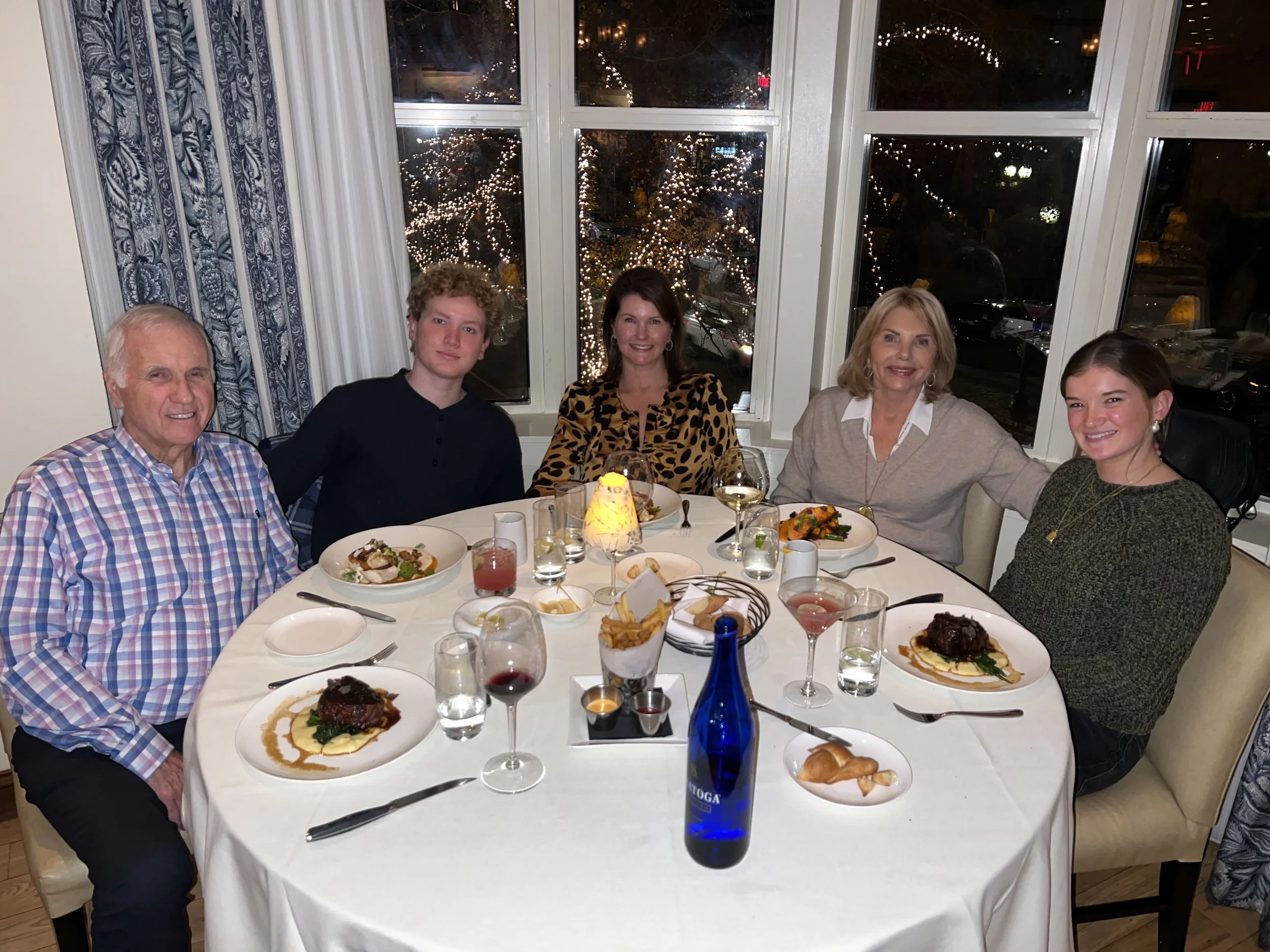 Six people sitting around a dining table with wine glasses, plates of food, and a blue bottle, inside a restaurant with large windows showing a decorated outdoor Christmas scene with trees and lights.