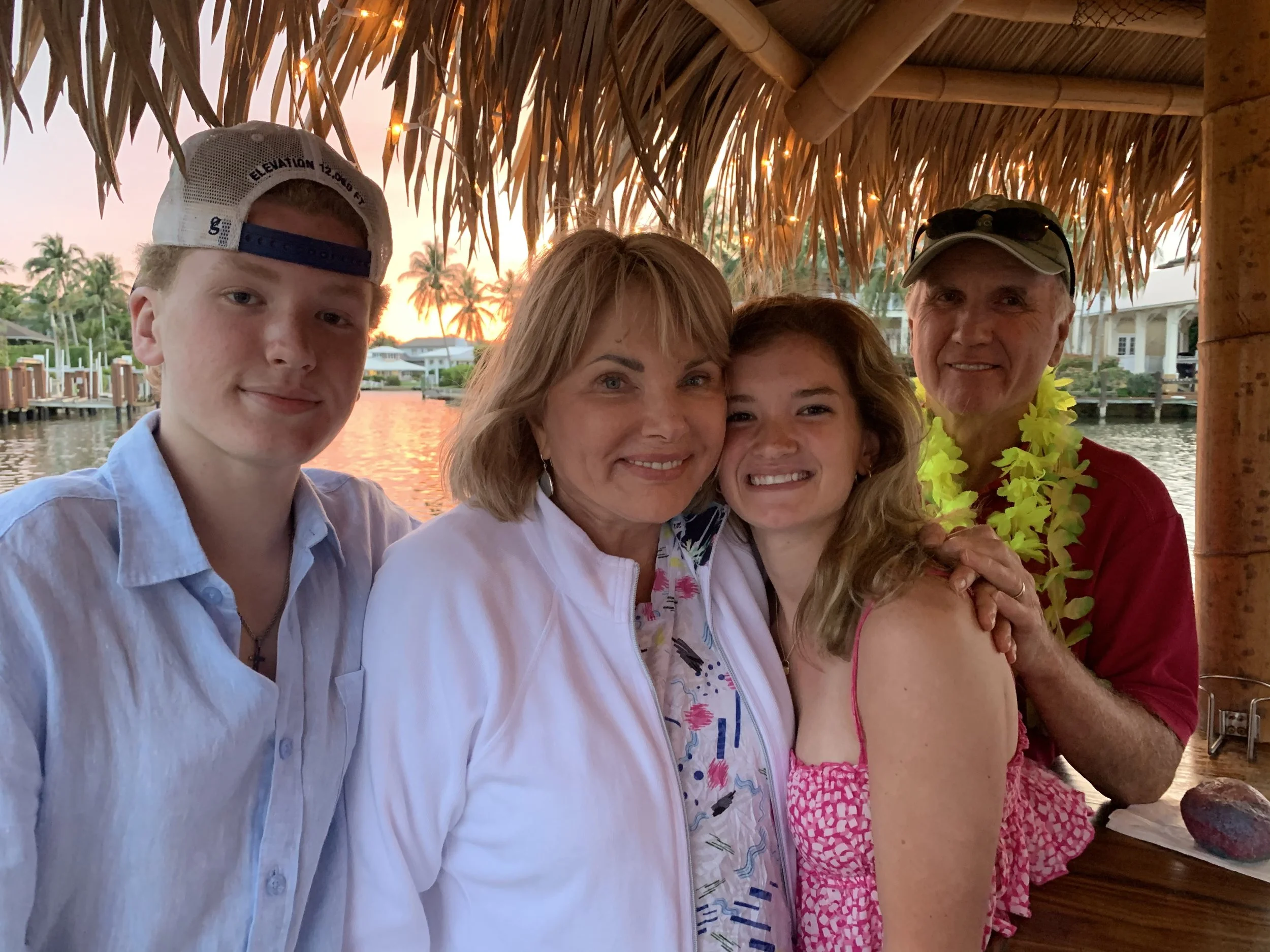 A family of five enjoying a tropical sunset at an outdoor tiki hut by water, smiling and posing for a photo.
