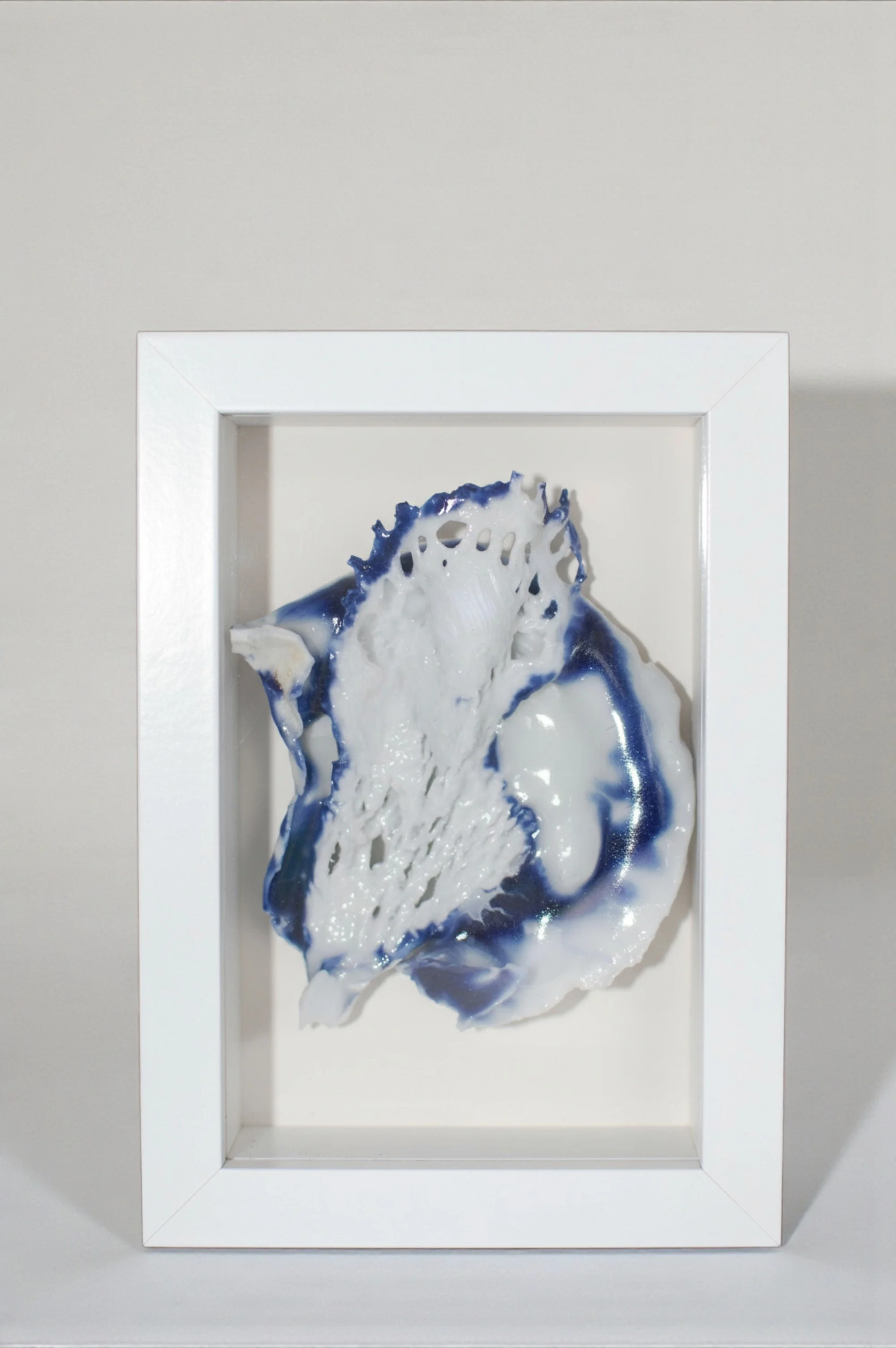 Une sculpture en céramique ou en peinture 3D blanche et bleue dans une vitrine en cadre blanc, représentant une forme organique ou abstraite.