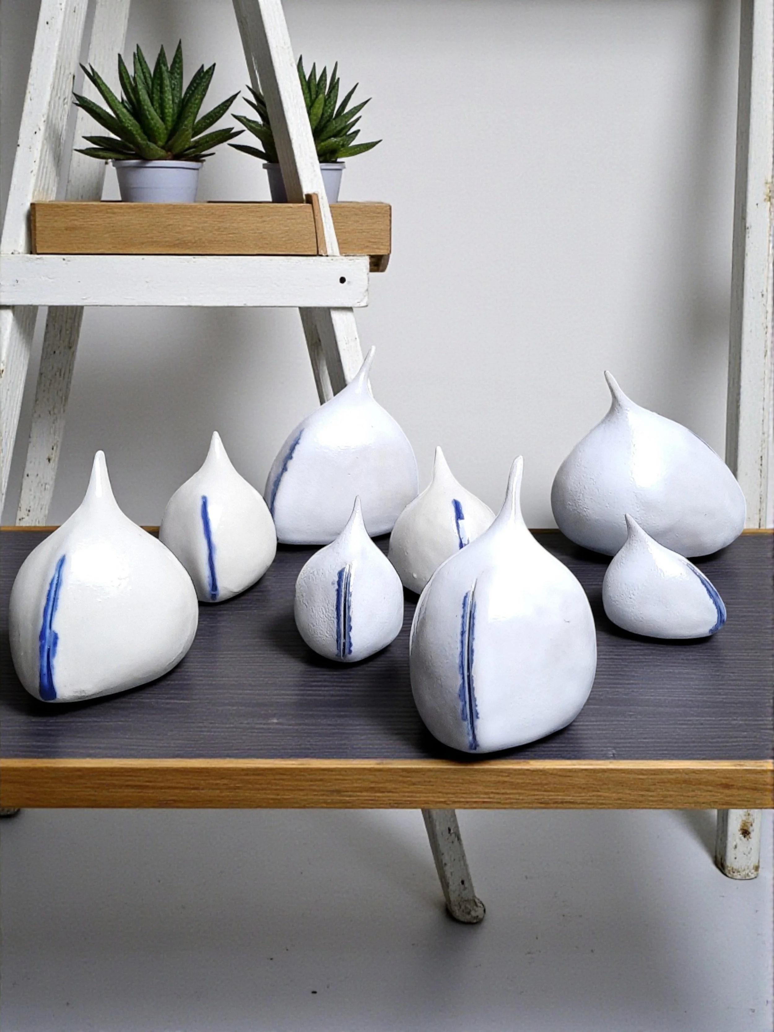 Une collection de sculptures en céramique blanches avec des lignes bleues, disposées sur une surface en bois, avec un étagère contenant des plantes succulentes en arrière-plan.