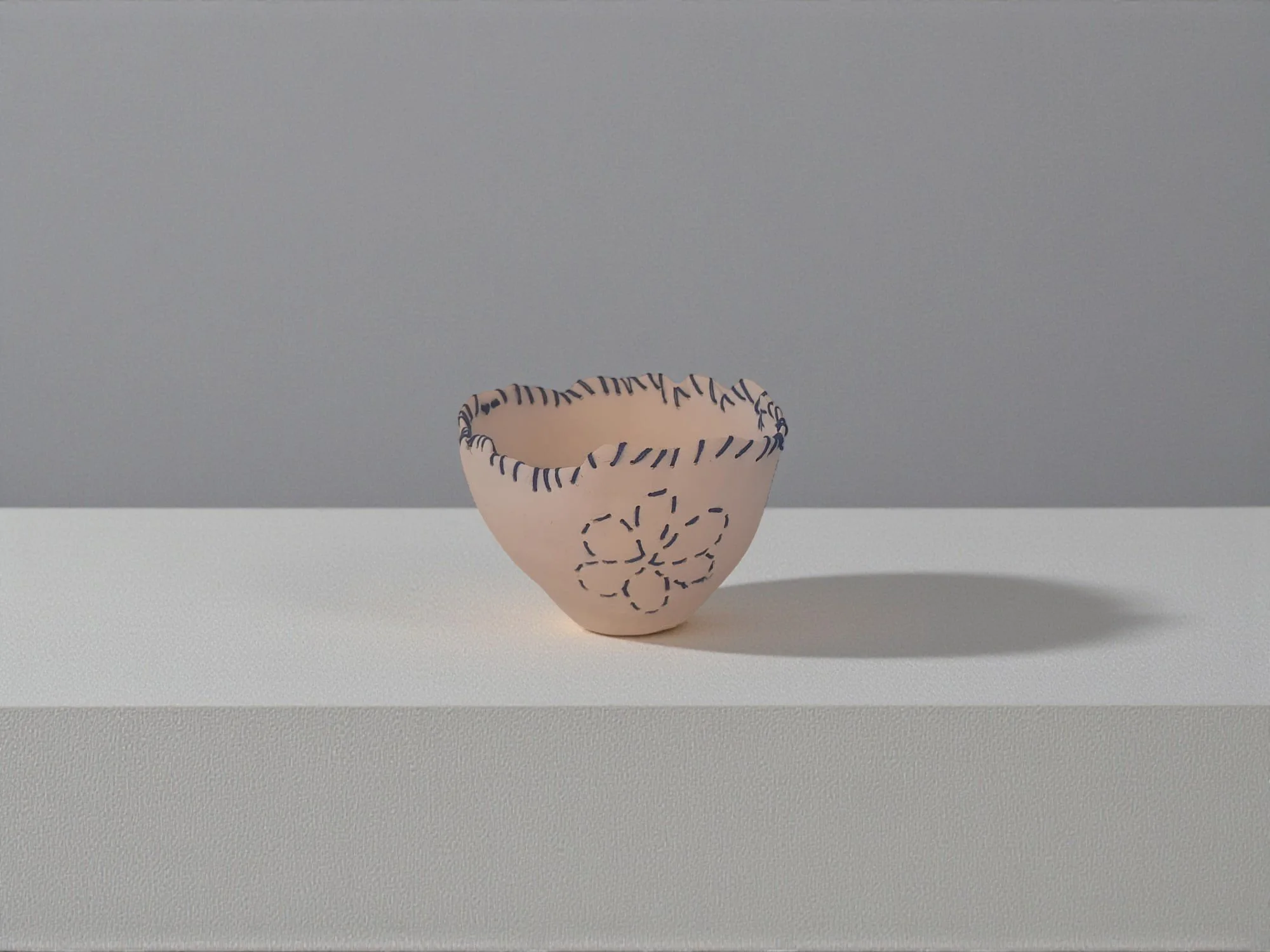 Une petite coupe en céramique blanche avec des motifs de broderie bleue, placée sur une surface blanche avec un fond gris. L'objet a un ombrage sur la droite.