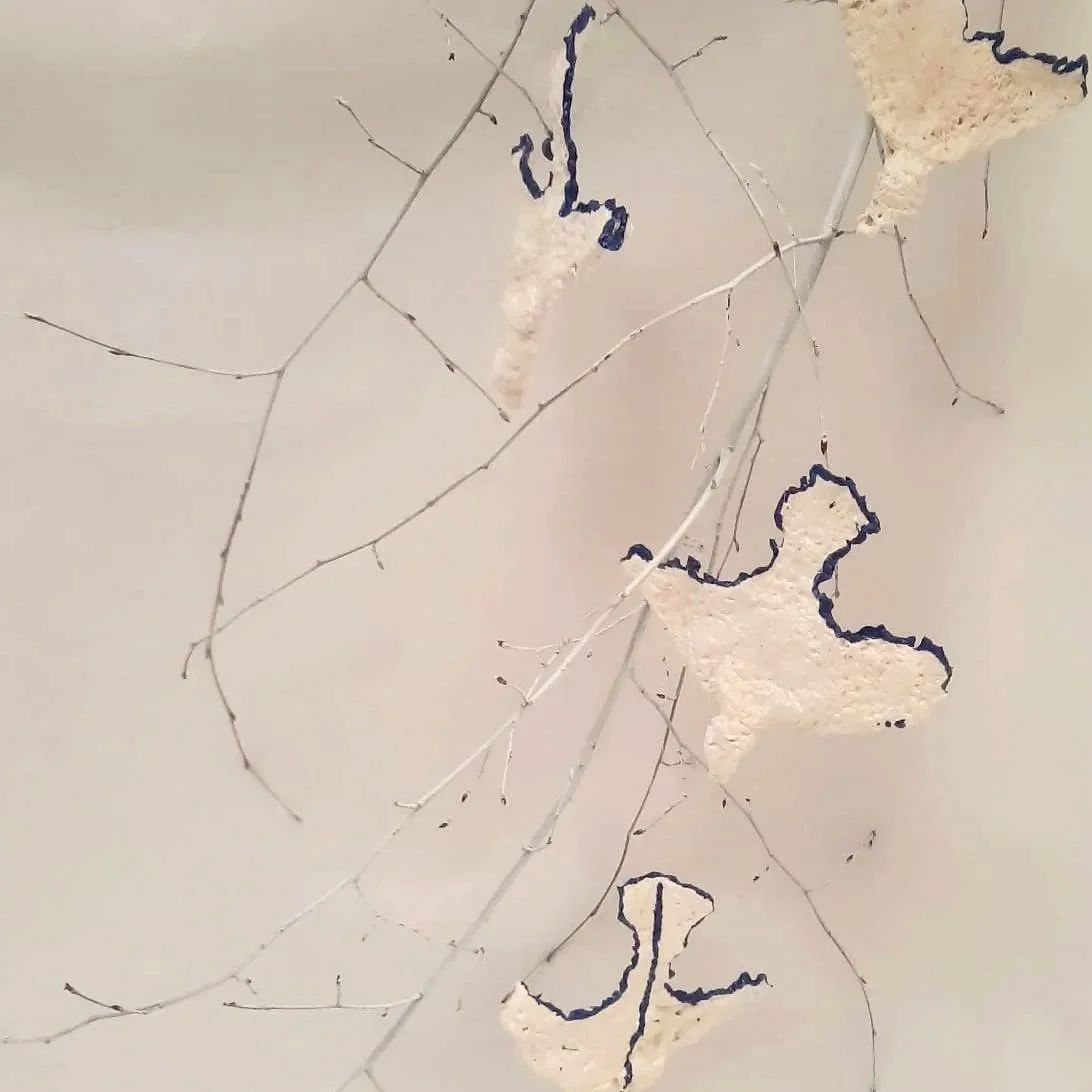 Une installation artistique composée de branches fines suspendues avec des morceaux de styromousse ou un matériau similaire, décorés de traits noirs évoquant des formes abstraites ou des animaux stylisés.