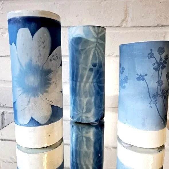 Trois lampes en verre avec des motifs floraux bleus et blancs, disposées sur une surface reflective devant un mur en briques blanches.